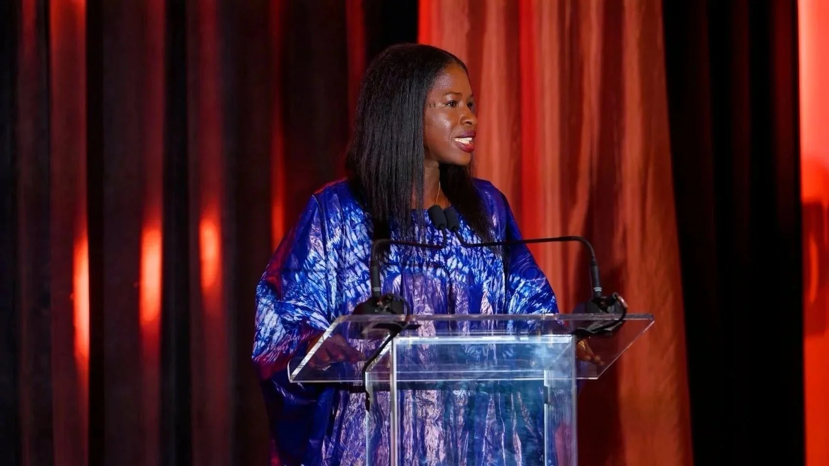A woman standing at a transparent podium with a microphone, speaking at an event with orange and black curtains in the background.