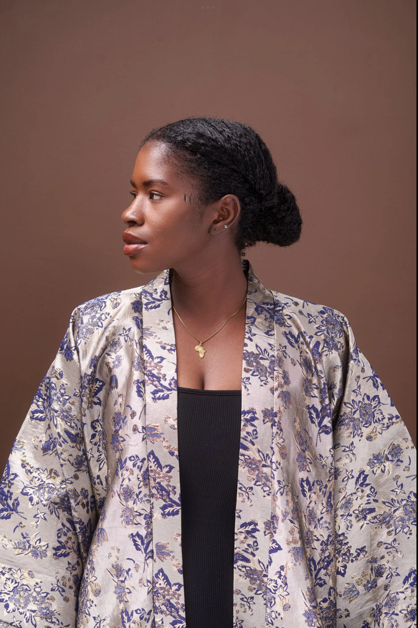 A woman with dark skin and black hair tied in a bun, wearing a floral blazer over a black top, gold necklace with an Africa-shaped pendant, earrings, and a small face tattoo, against a plain brown background.