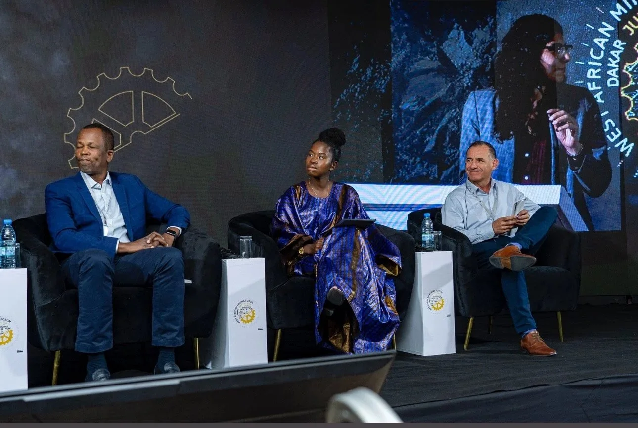 Three people sitting on stage participating in a panel discussion at a conference, with a large screen behind showing a woman speaking.