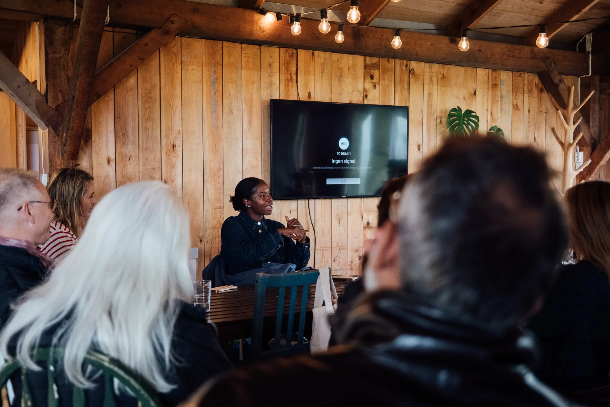 A group of people gathered in a rustic wooden room, attending a presentation or meeting. A woman is speaking, seated at a table in front of a large flat screen television displaying messages in Norwegian indicating no signal. The room is decorated wi