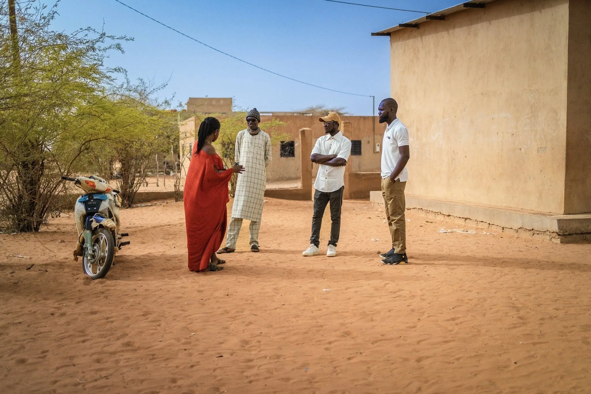 Four people standing outdoors on a reddish dirt ground in front of a building with a yellowish wall, some trees, and a clear blue sky. One person is wearing a red dress, while the others are dressed casually in shirts and pants. There is a motor scoo