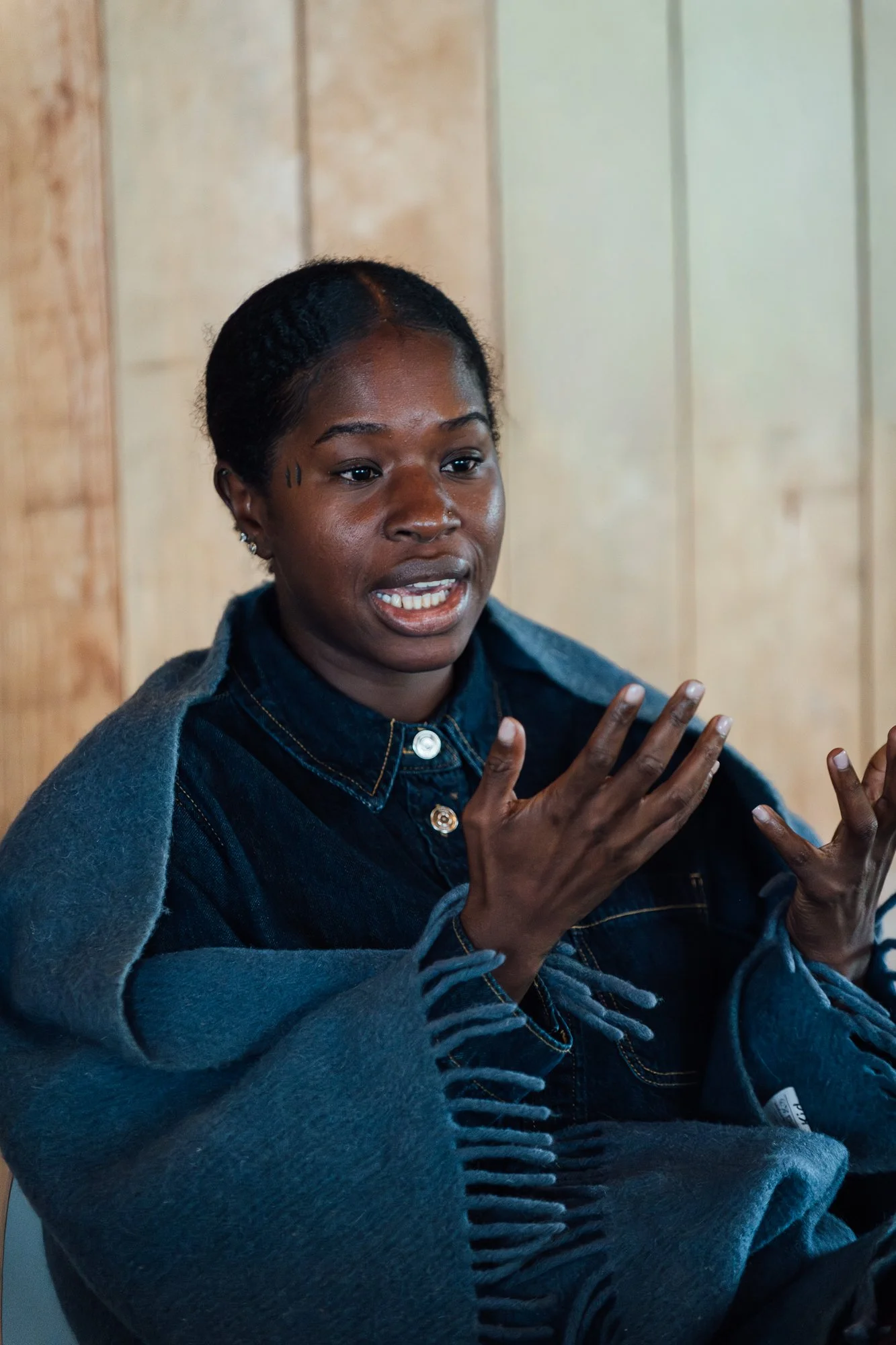 A woman with dark skin and short black hair, speaking and gesturing with her hands, wearing a denim jacket and a grey sweater, sitting in front of a wooden wall.