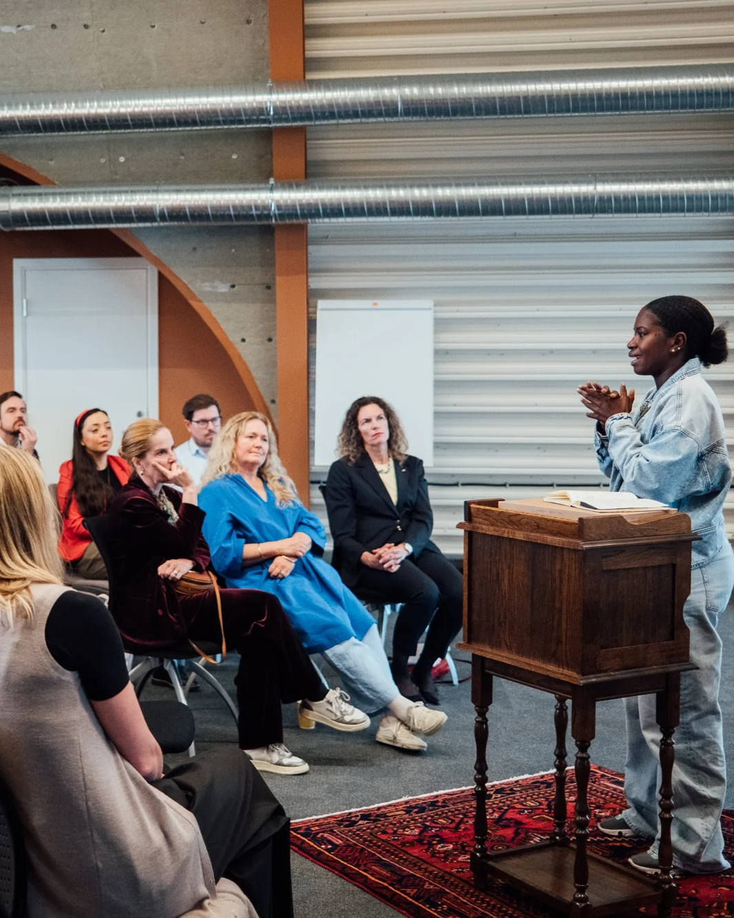 A woman speaking in front of a seated audience inside a room with exposed ductwork and metal siding. The speaker stands behind a wooden podium, and the audience listens attentively.