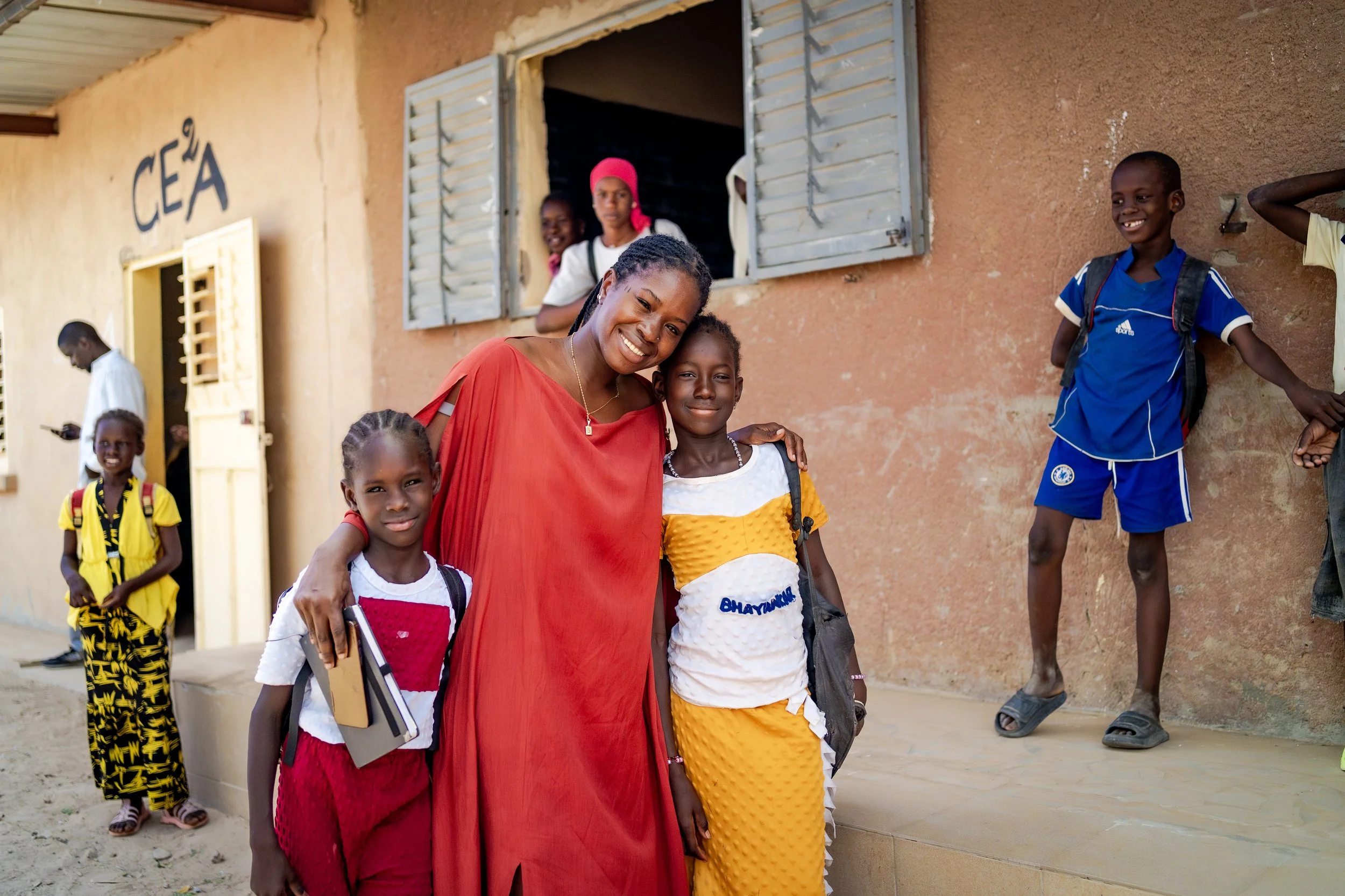 A woman and three children standing outside a school building with a sign that reads 'CEA'. The woman is smiling and wearing a red dress, with her arm around one child. The children are smiling, carrying backpacks, and standing around the woman.
