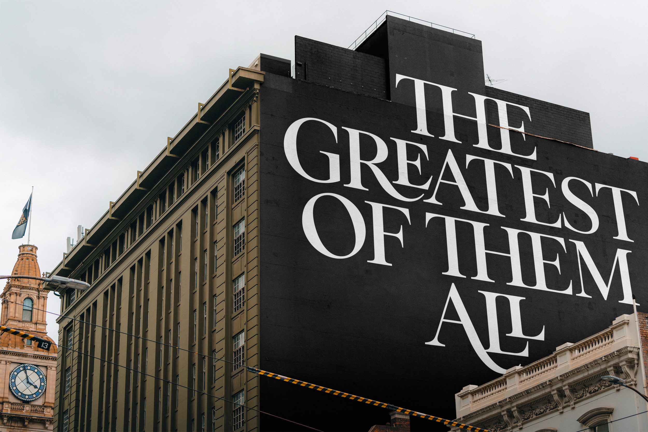A large black billboard on the side of a building with white text stating, 'The Greatest of Them All' in a stylized font. The surrounding area includes a historic beige building with a clock and a flag.