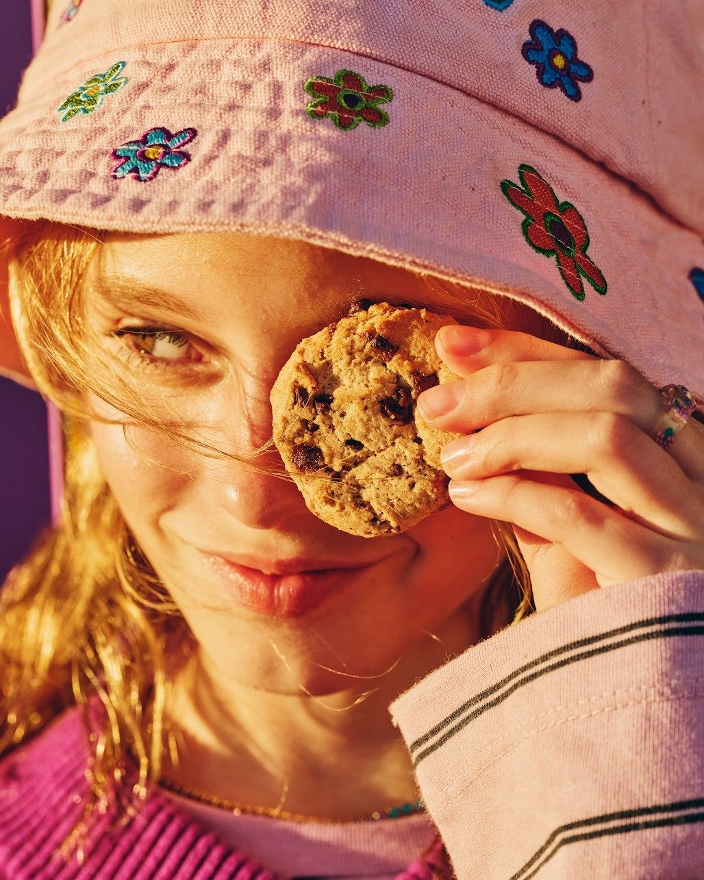 A young woman with red hair wearing a pink hat with embroidered colorful flowers and a pink sweater, holding a chocolate chip cookie next to her eye in an outdoor setting during sunset.