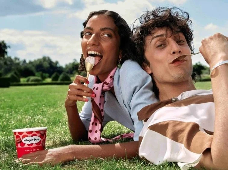 Two people lying on the grass outdoors, enjoying ice cream, with a container of Haagen-Dazs ice cream beside them.