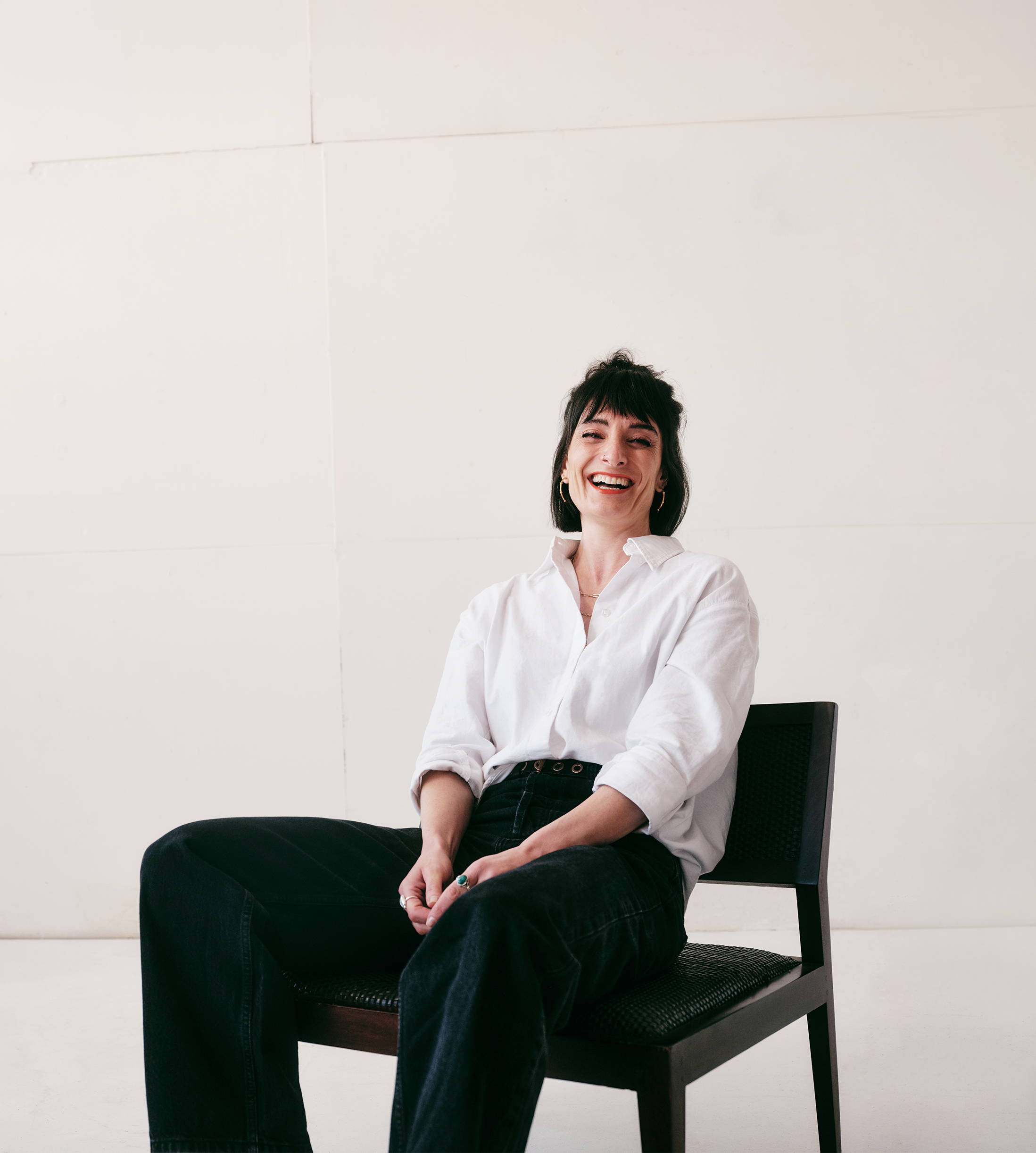 A woman with short dark hair and gold hoop earrings, wearing a white shirt and black pants, sitting on a black chair against a plain white wall, smiling and laughing.