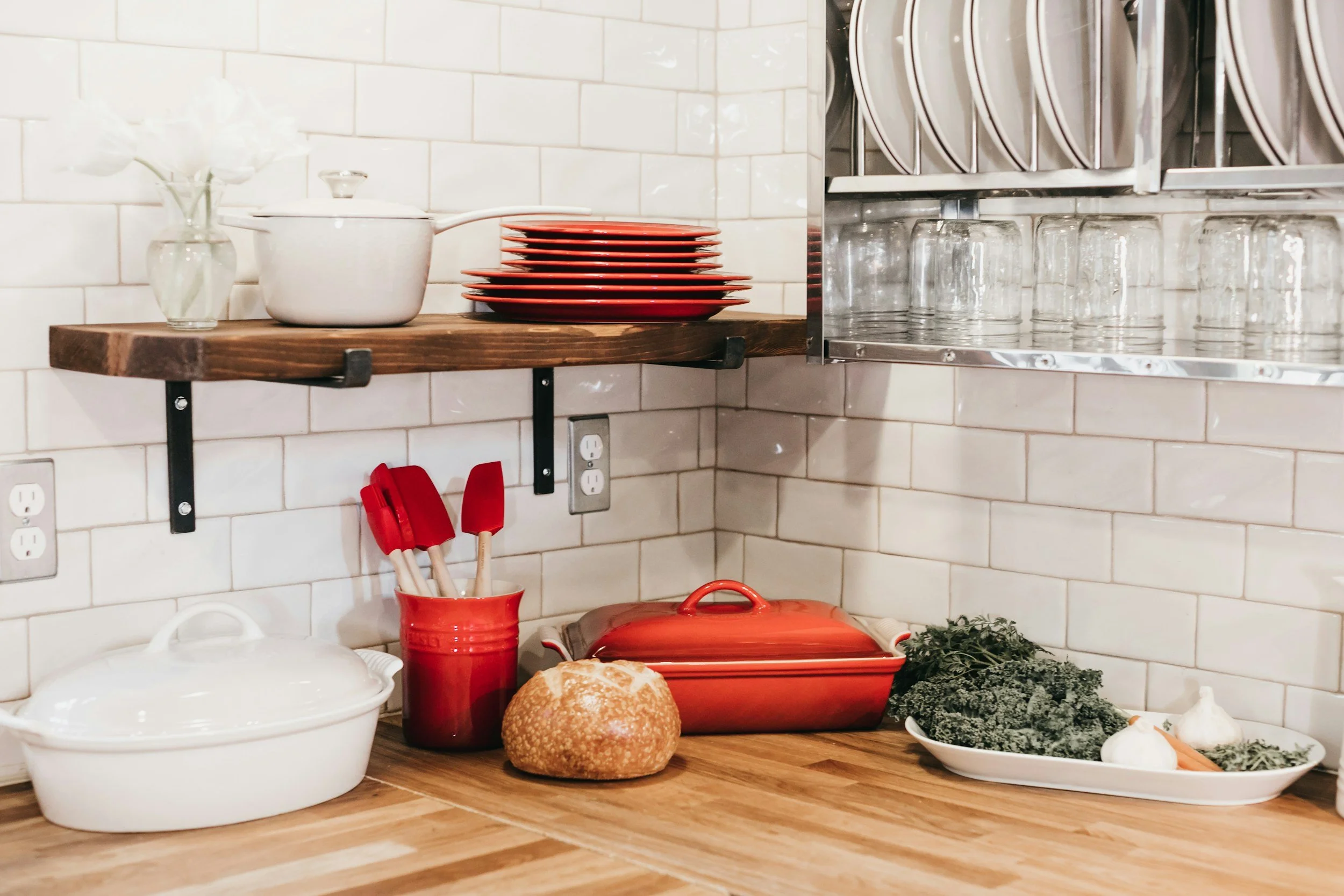 Kitchen countertop with white ceramic dish casserole, red utensils container with spatula, bread, red baking dish, garlic, vegetables, and greens. White subway tile backsplash, wooden shelf with red plates, and glasses stored above.
