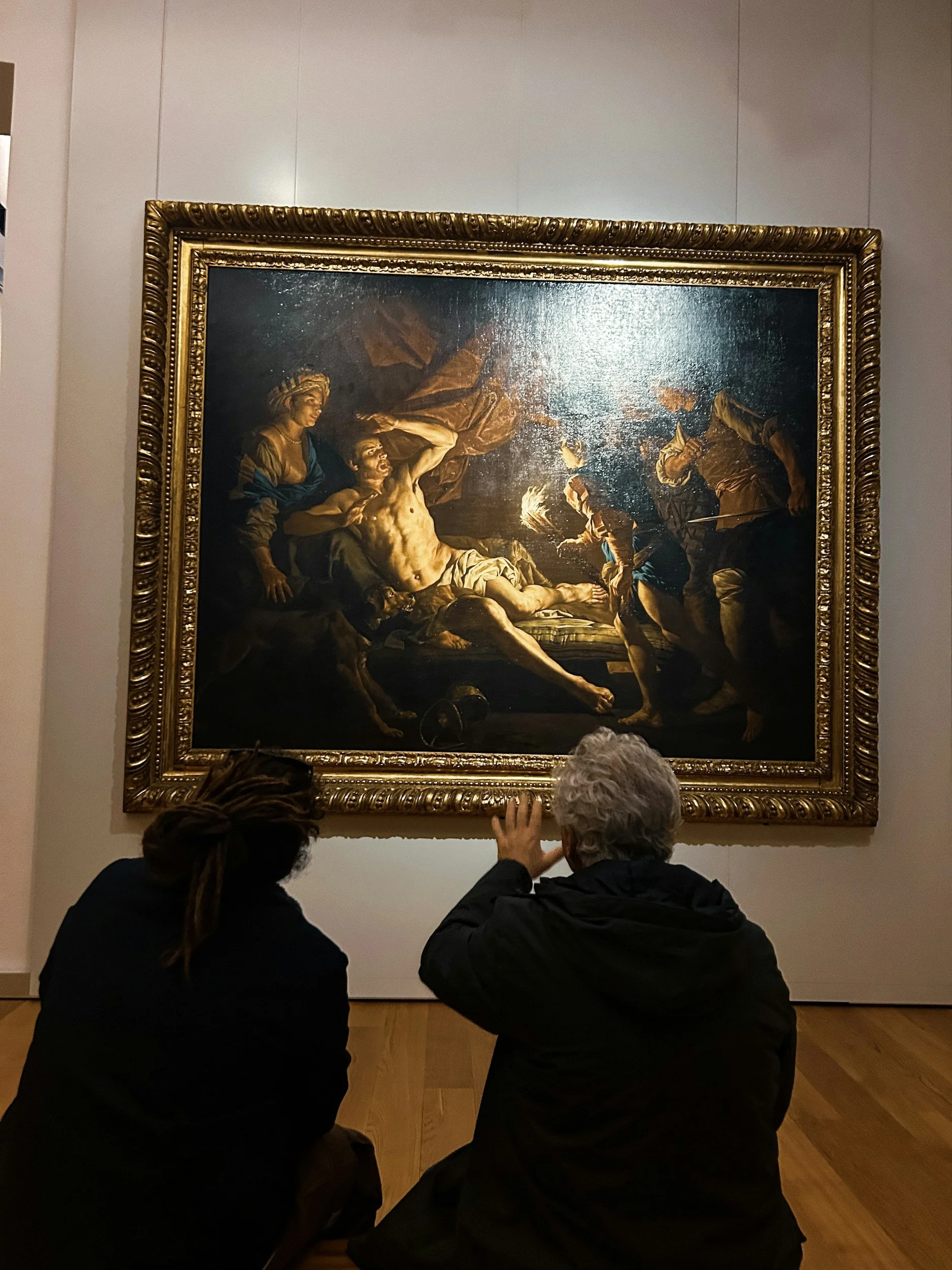 Two people sitting on the floor looking at a large classical painting in a museum.