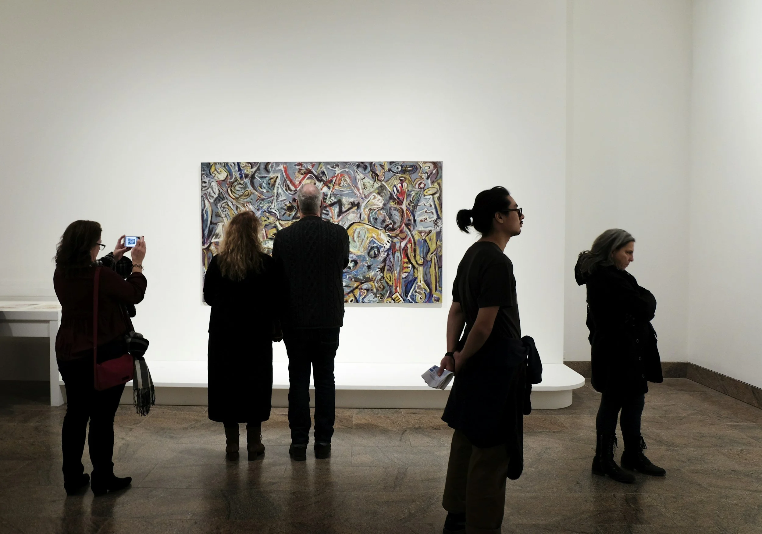 Multiple people viewing and photographing an abstract painting in a museum gallery. Five individuals are visible, with some observing the artwork and others taking photos. The painting features colorful, dynamic abstract shapes and lines.