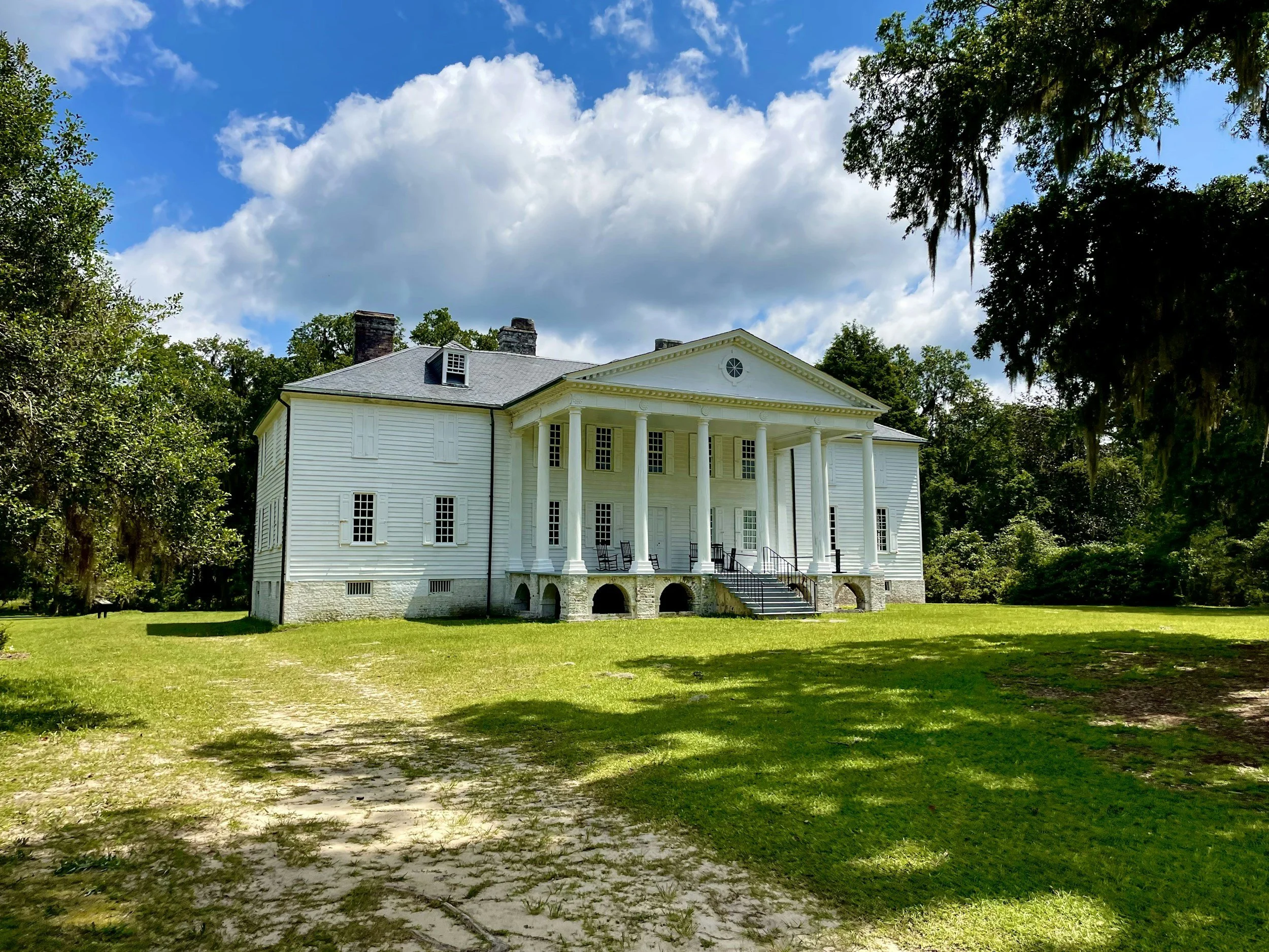White historic mansion with columns on a grassy lawn under a partly cloudy sky.
