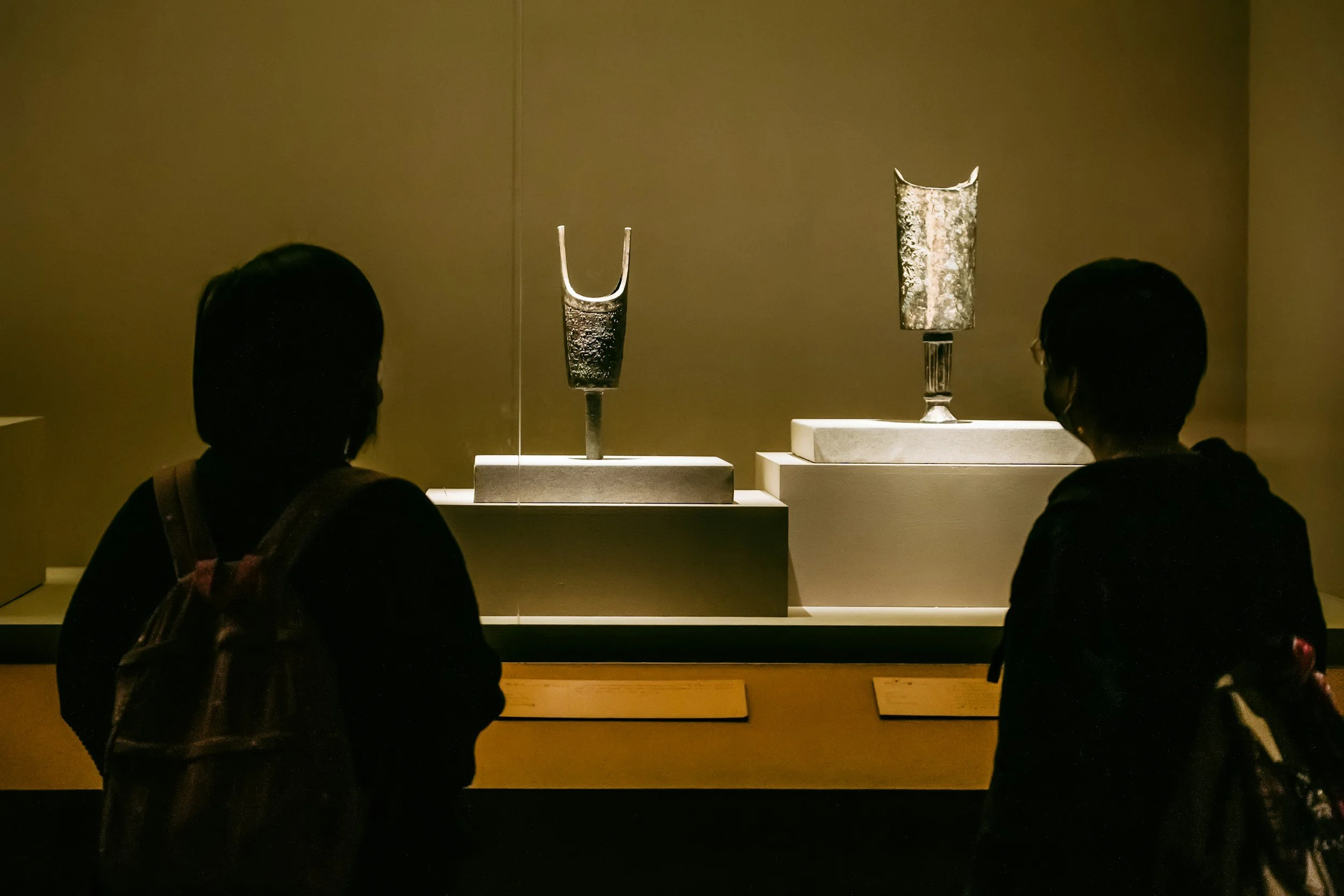 Two people observing ancient artifacts in a museum display case with dim lighting.