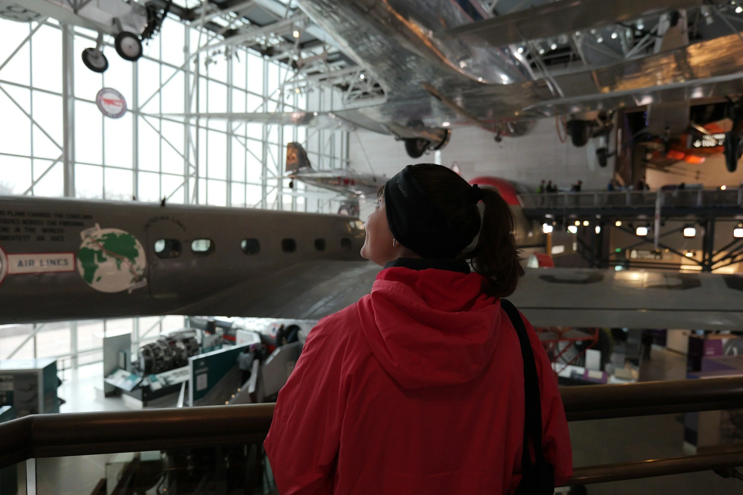 A woman wearing a red jacket and black headband looking at vintage aircraft inside an airport or hangar with large glass windows and metal framework.