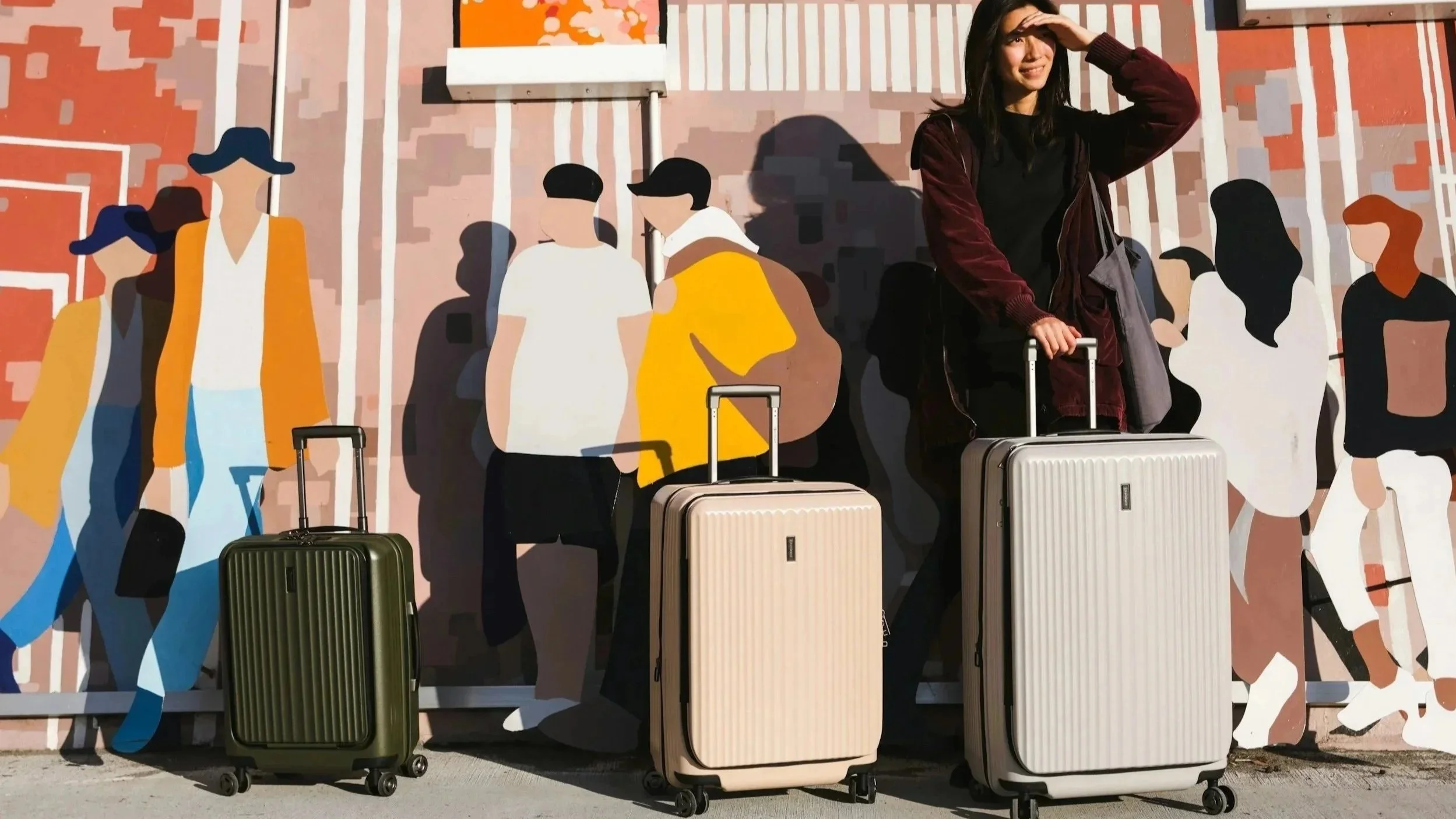 A woman standing outside an airport holding a suitcase, with a colorful mural of abstract human figures in the background.
