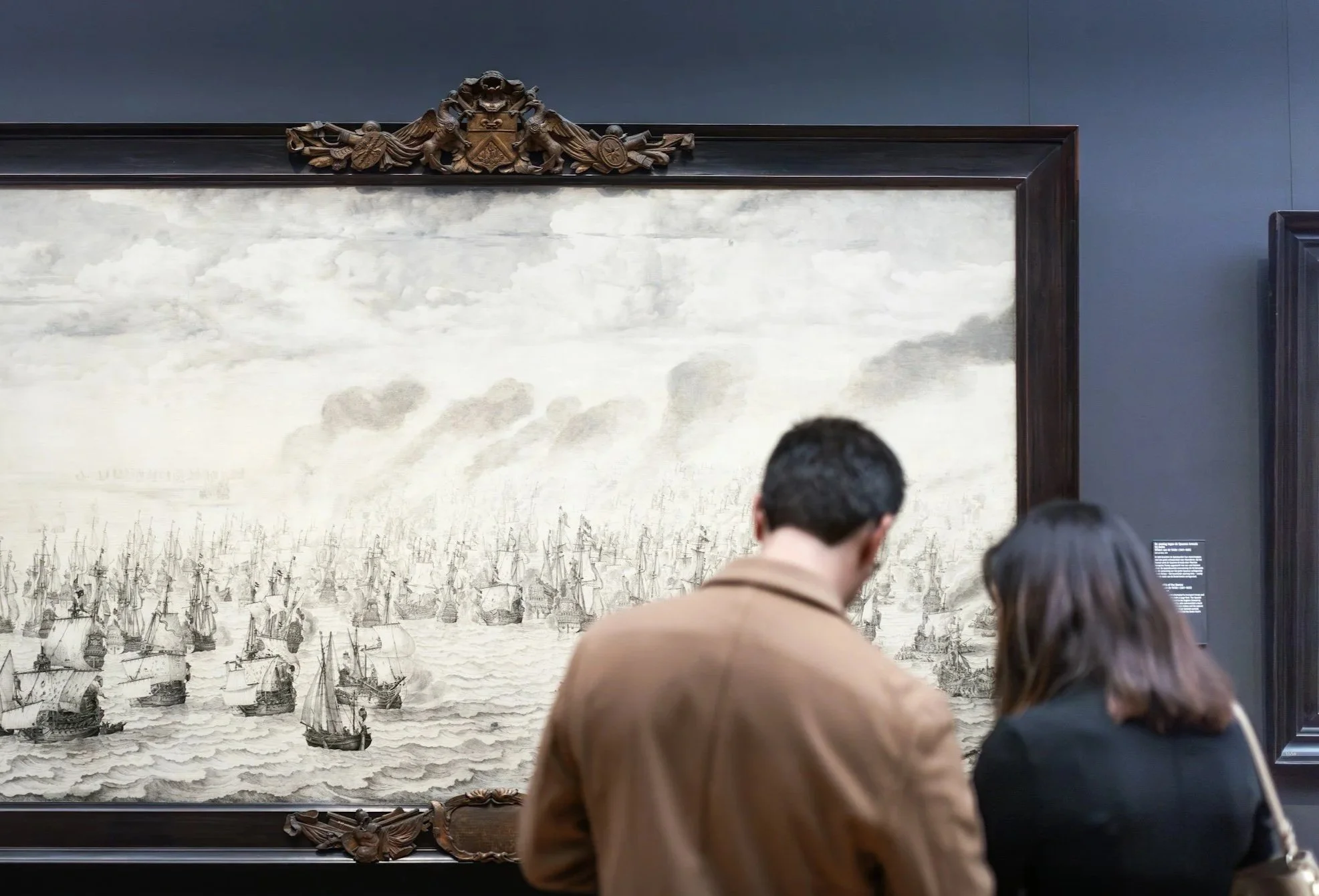 People viewing a historical painting of sailing ships at a museum.