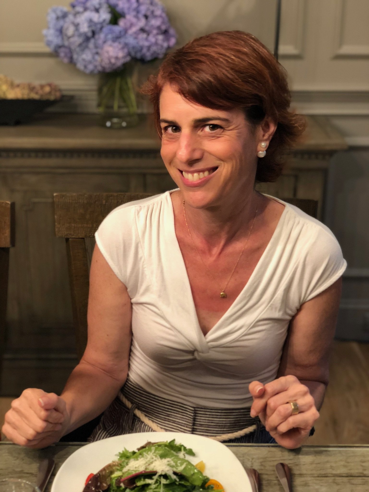 A woman with short reddish-brown hair, wearing a white top, smiling while sitting at a table with a plate of salad in front of her. In the background, there are purple flowers on a cabinet.