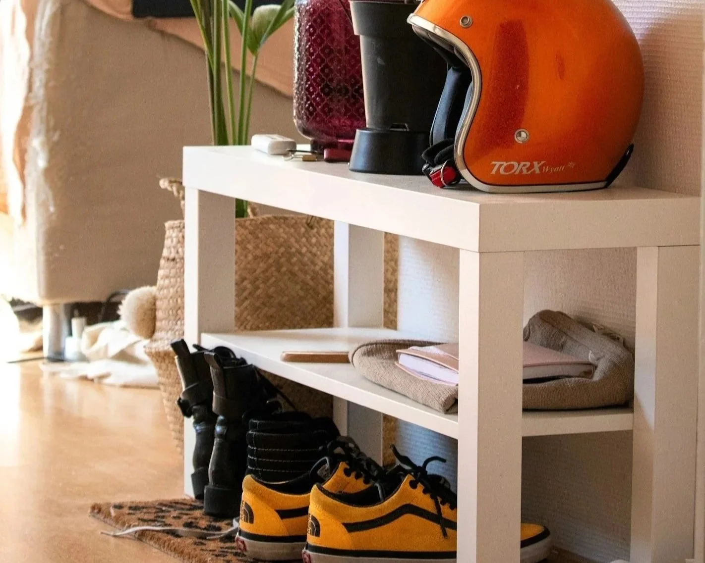 A white shoe rack with three shelves containing a pair of yellow and black sneakers, a pair of black boots, and a pair of tan shoes. On top of the rack, there are a red helmet, a black helmet, a white charger, and a red glass vase with green leaves inside. In the background, there is a beige sofa, a woven basket, and other household items.