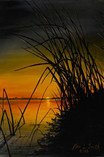 Silhouette of tall grass by a lake during sunset with an orange and yellow sky.