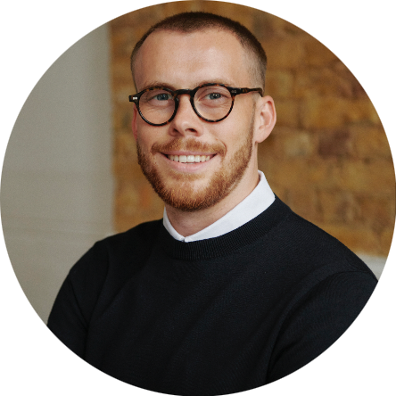 Portrait of a man with short hair, glasses, a beard, and a mustache, wearing a black sweater over a collared shirt, smiling in front of a brick wall.