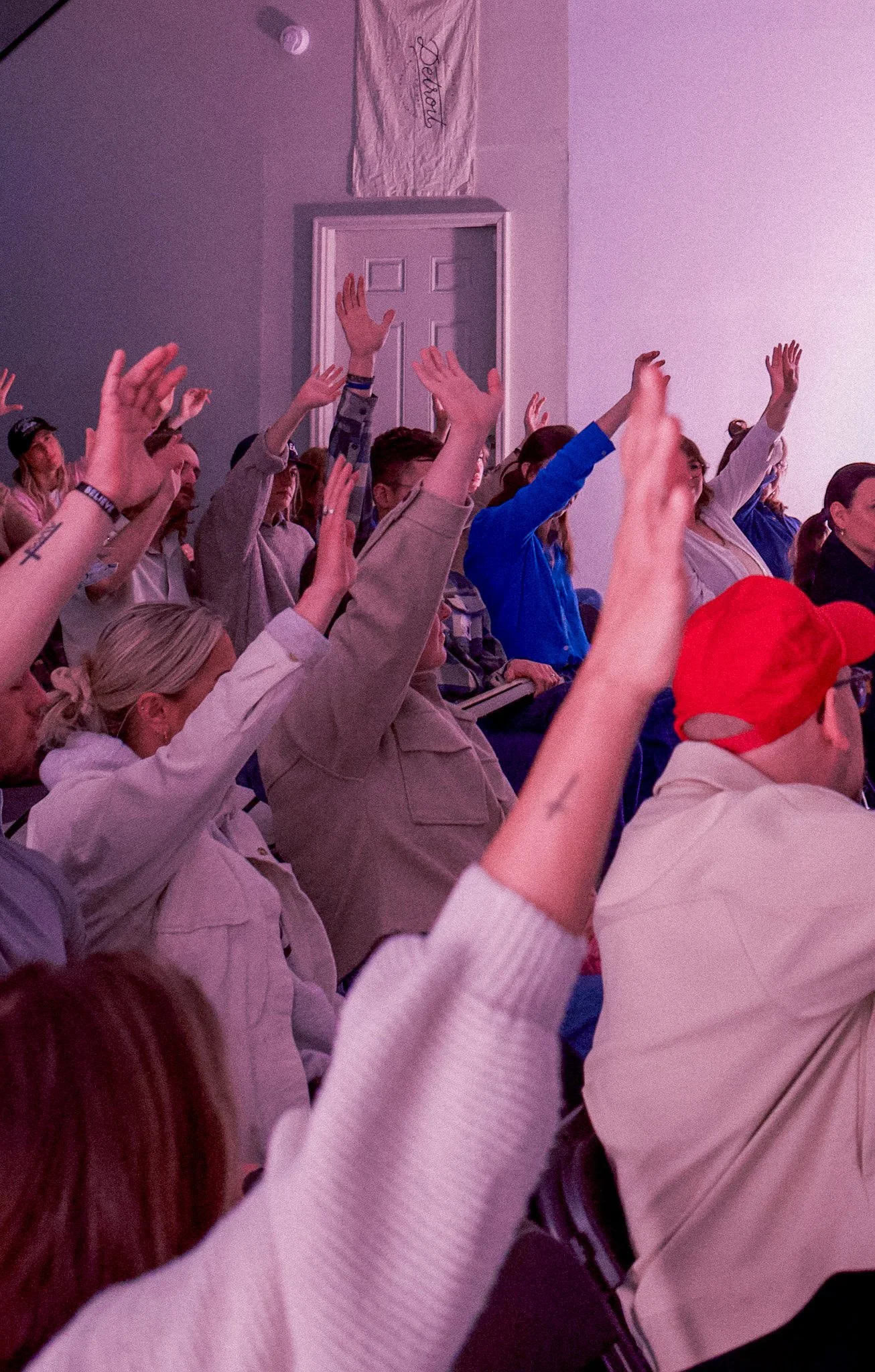 Audience members raising their hands in a large indoor gathering with a brightly lit theater or conference room.