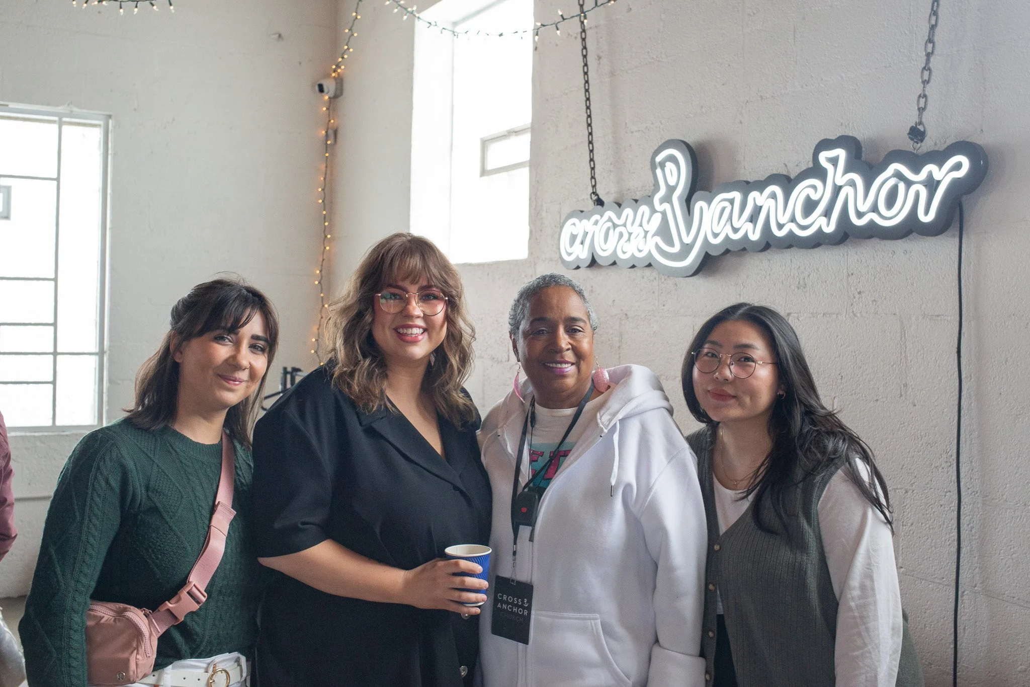 Four women standing together smiling at an event called Cross Anchor. They are indoors against a white brick wall with a neon sign that says 'Cross Anchor' and some string lights.