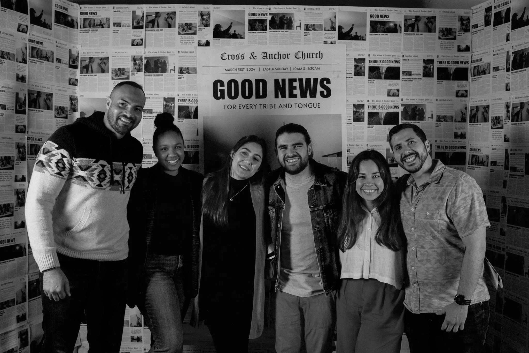A group of six smiling people standing closely together in front of a large sign that reads "Good News" at a church event, with walls covered in newspapers.