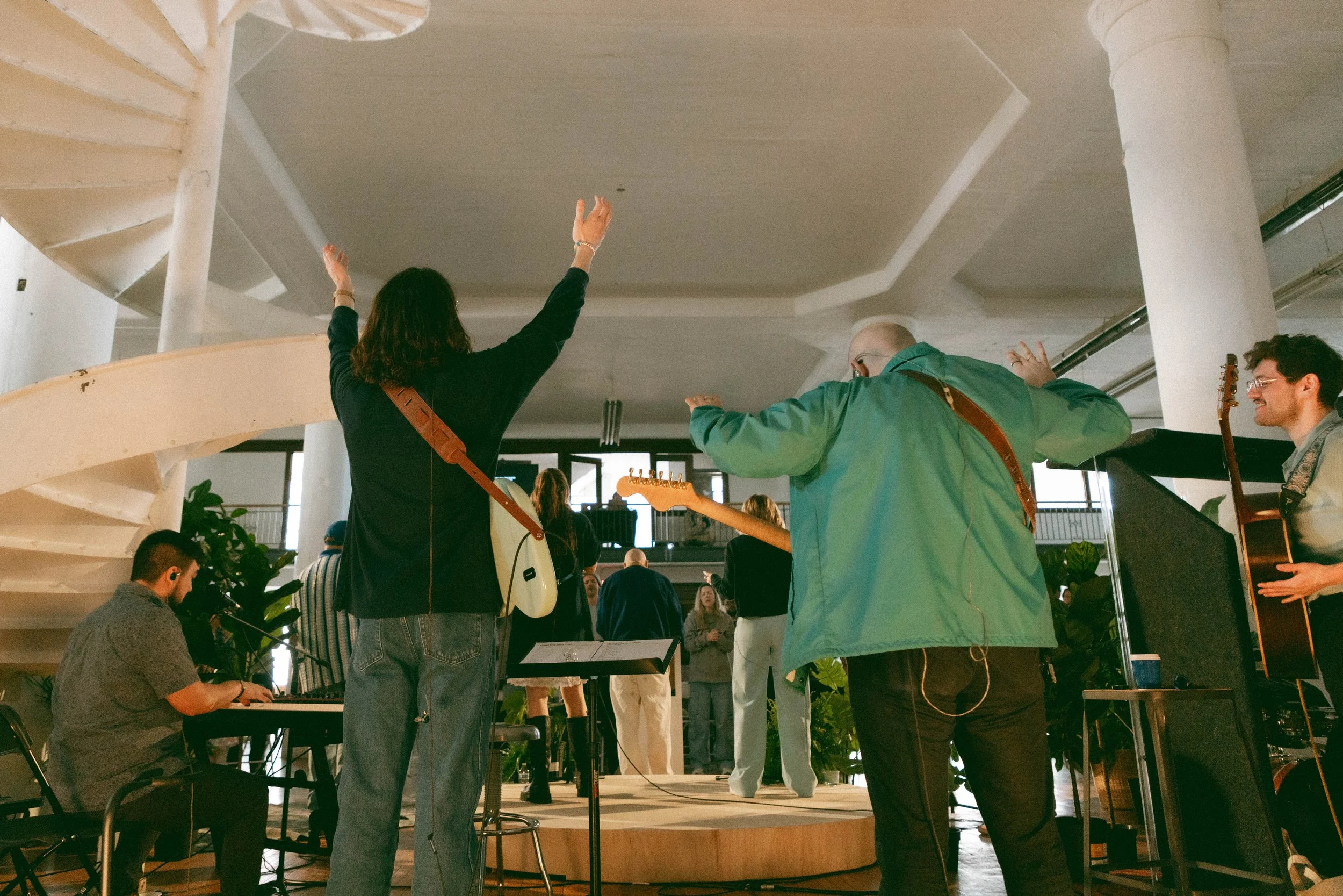 Group of musicians performing on stage, seen from behind, with one woman raising her hands and musicians holding guitars and a keyboard.