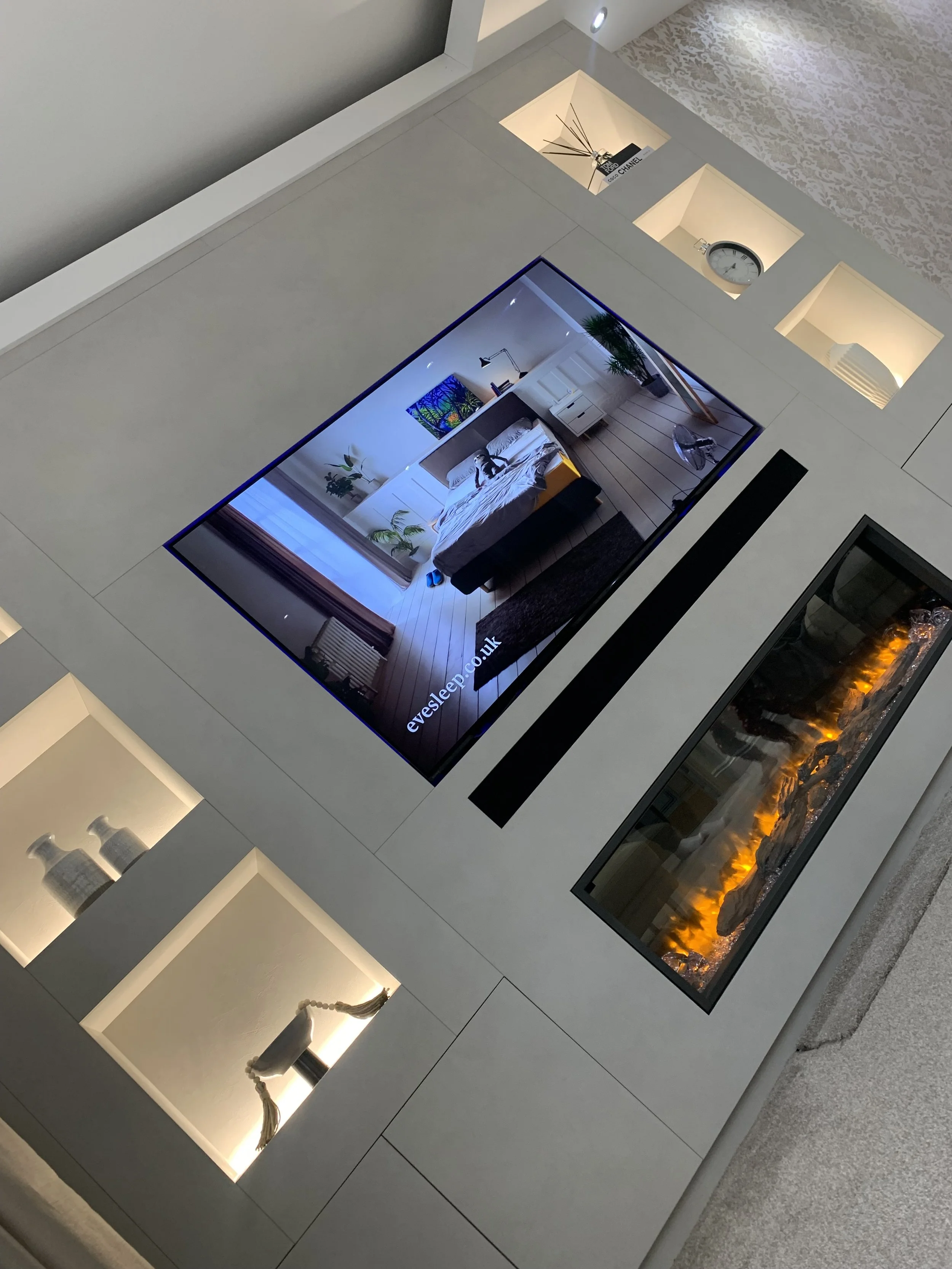 A modern, minimalist living room with a mounted flat-screen TV displaying a bedroom scene. The wall behind the TV has built-in illuminated shelves displaying decorative items and a clock. To the right of the TV, there is a long, rectangular fireplace