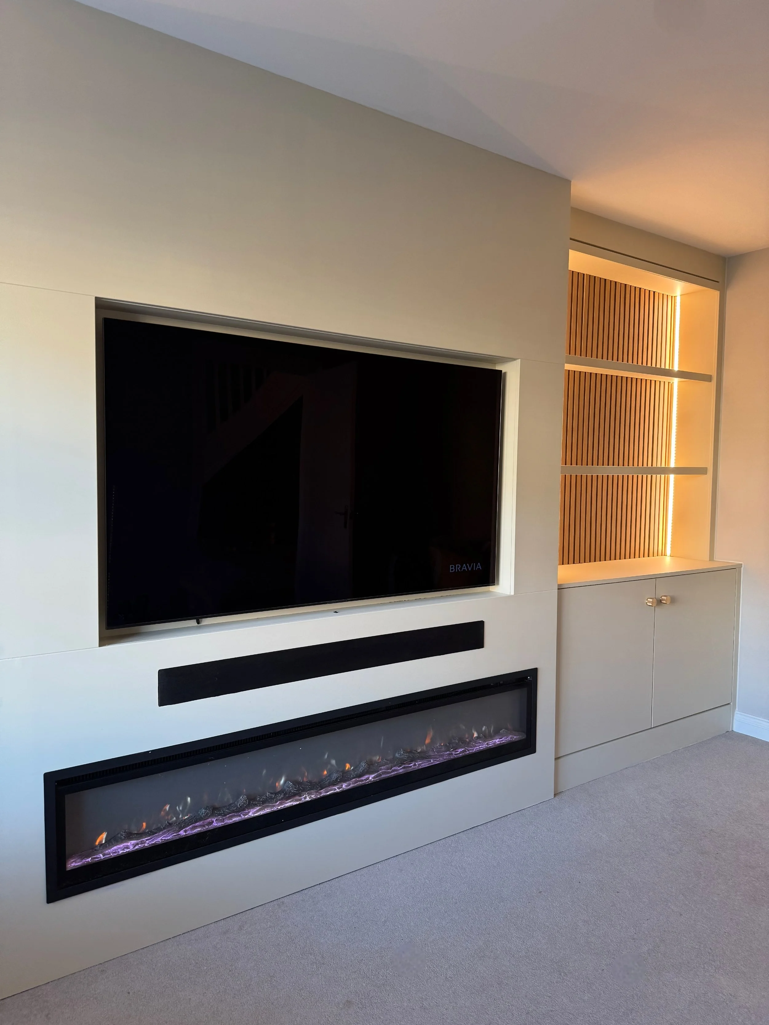 Living room wall with a mounted flat-screen TV, electric fireplace below, and a built-in wooden shelf with lighting to the right.