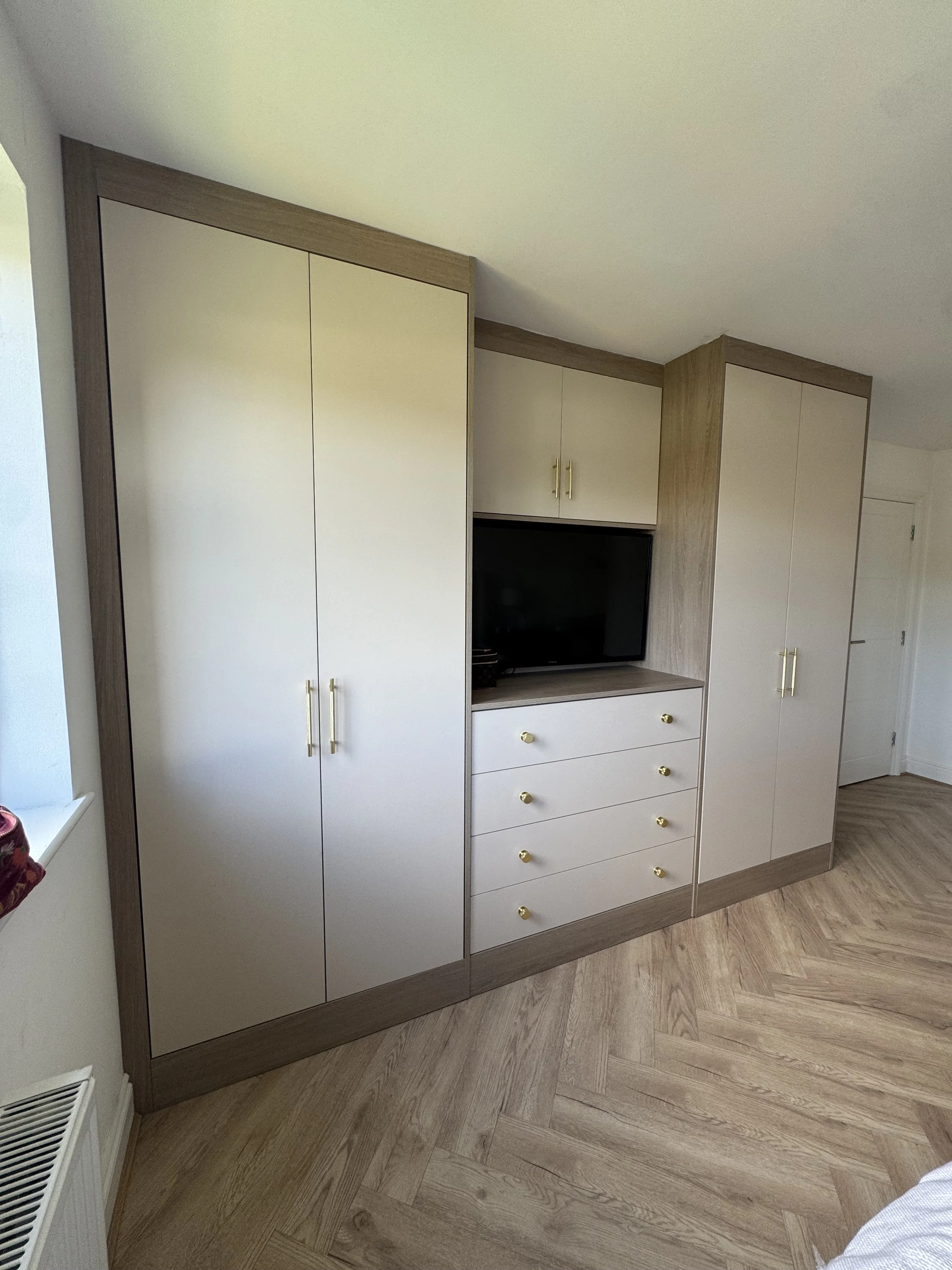 A built-in wardrobe with white doors, gold handles, and wood trim, containing cabinets, drawers, and a space for a television, set on a wood floor in a room with white walls.