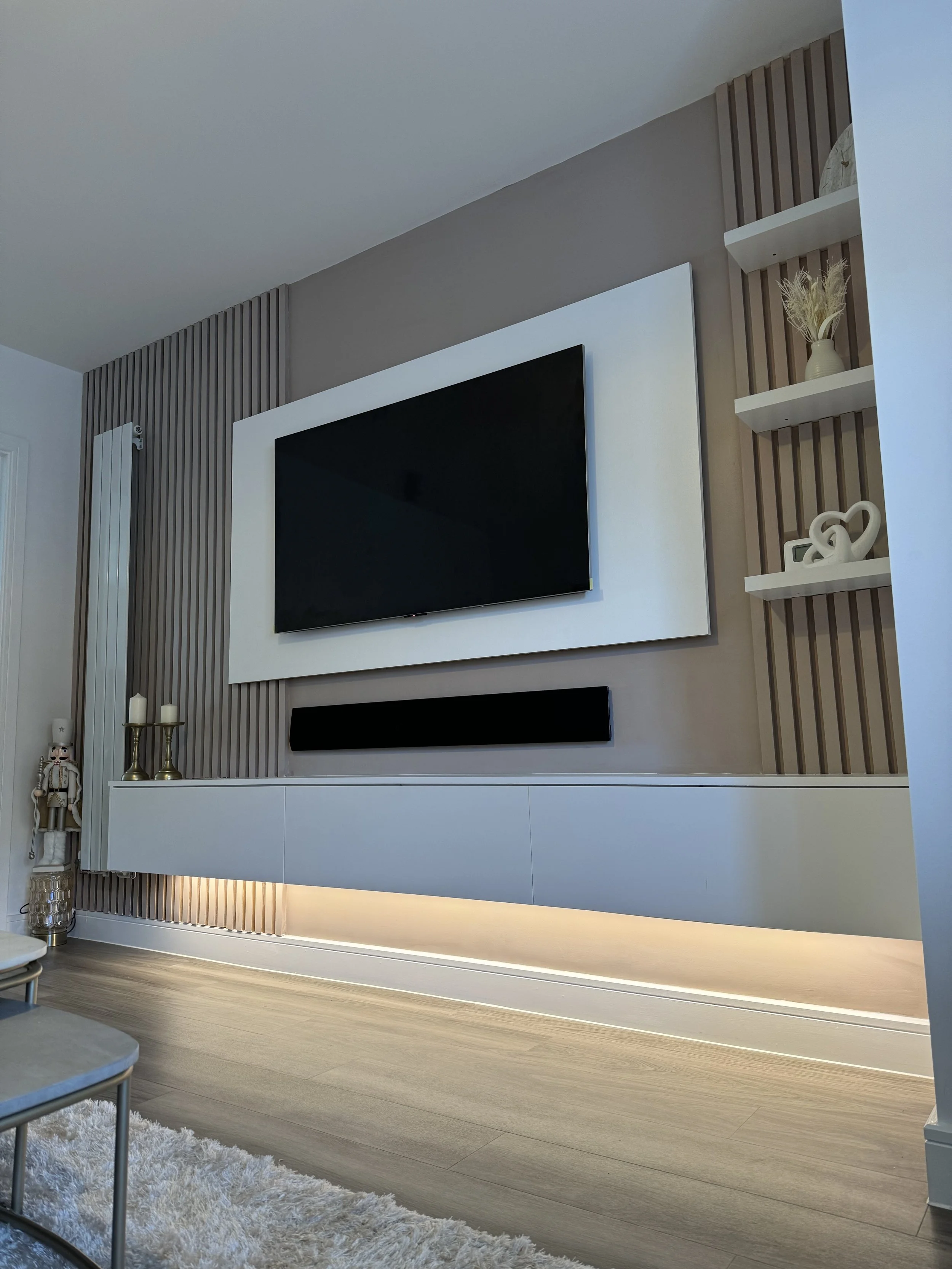 Modern living room wall with a large flat-screen TV, decorative white frame, black soundbar underneath, wooden slatted wall panel, white shelves with decorative items, candles, and a nutcracker, light wood flooring, and a beige shaggy rug.