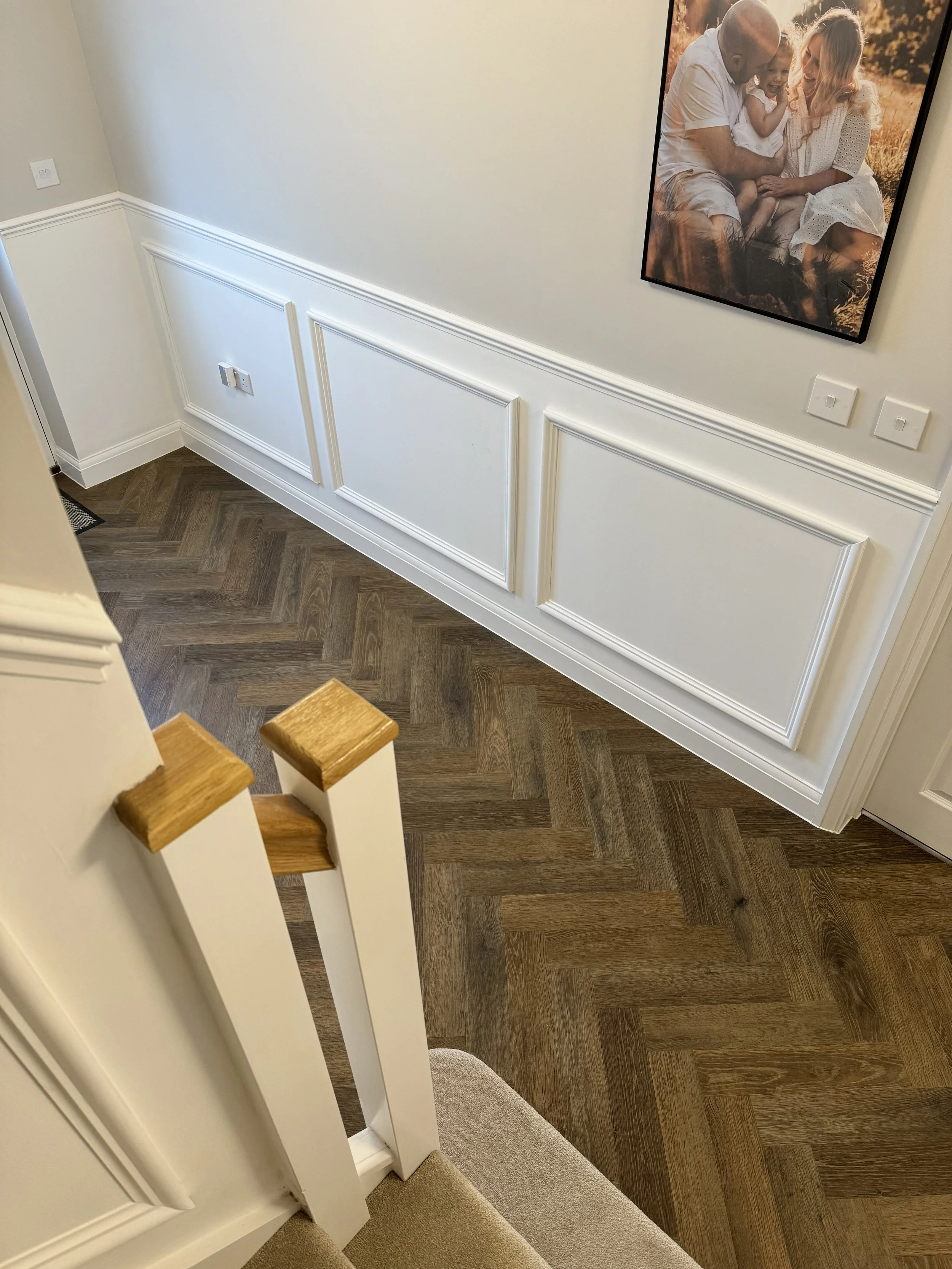Interior view of a hallway with wooden flooring, white wainscoting, a wall with a framed family photo, and a portion of a staircase with wooden handrails.