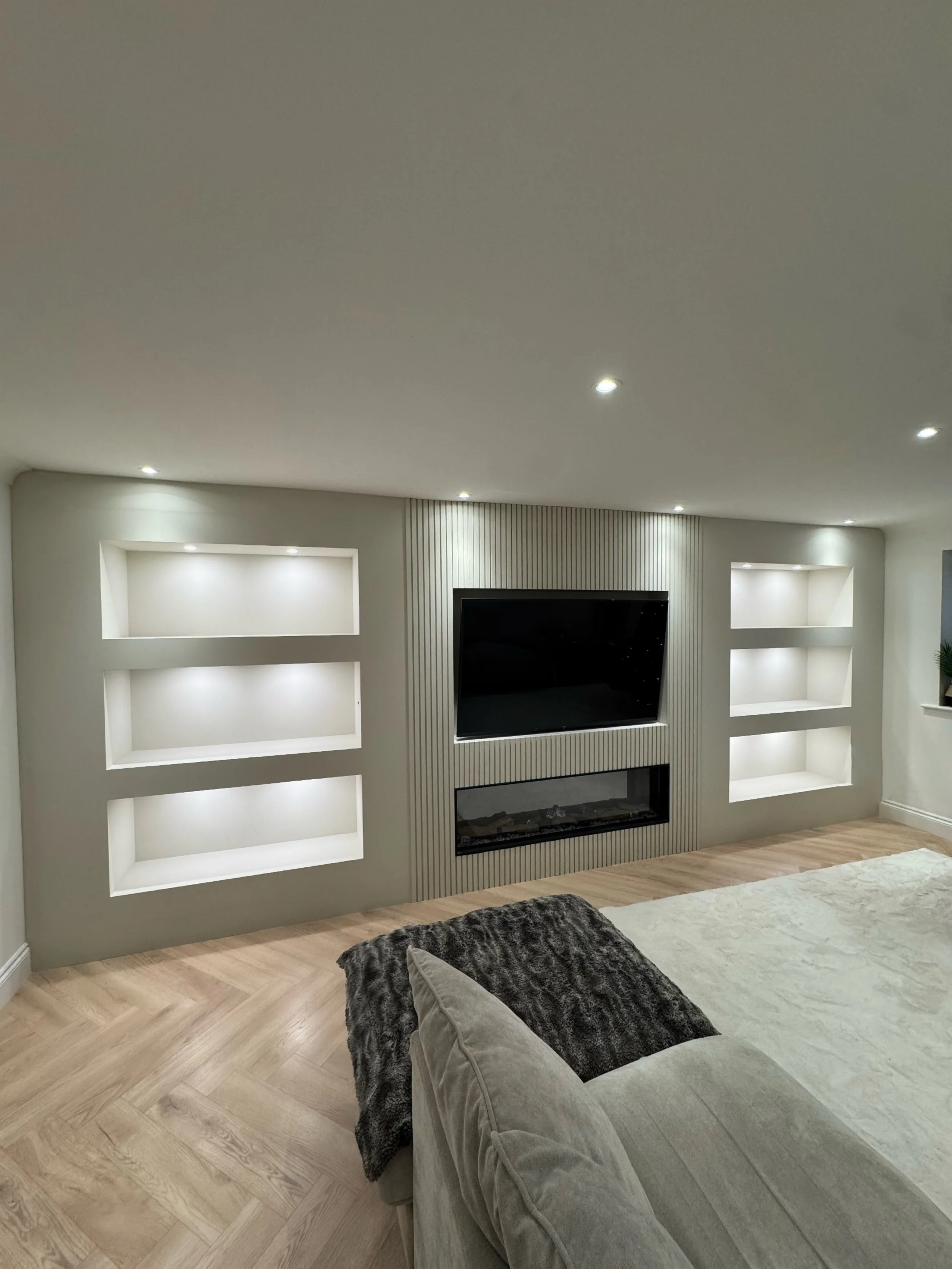 Living room with built-in wall shelves, a wall-mounted TV, a modern fireplace, a beige sofa with a cushion, and wood flooring.