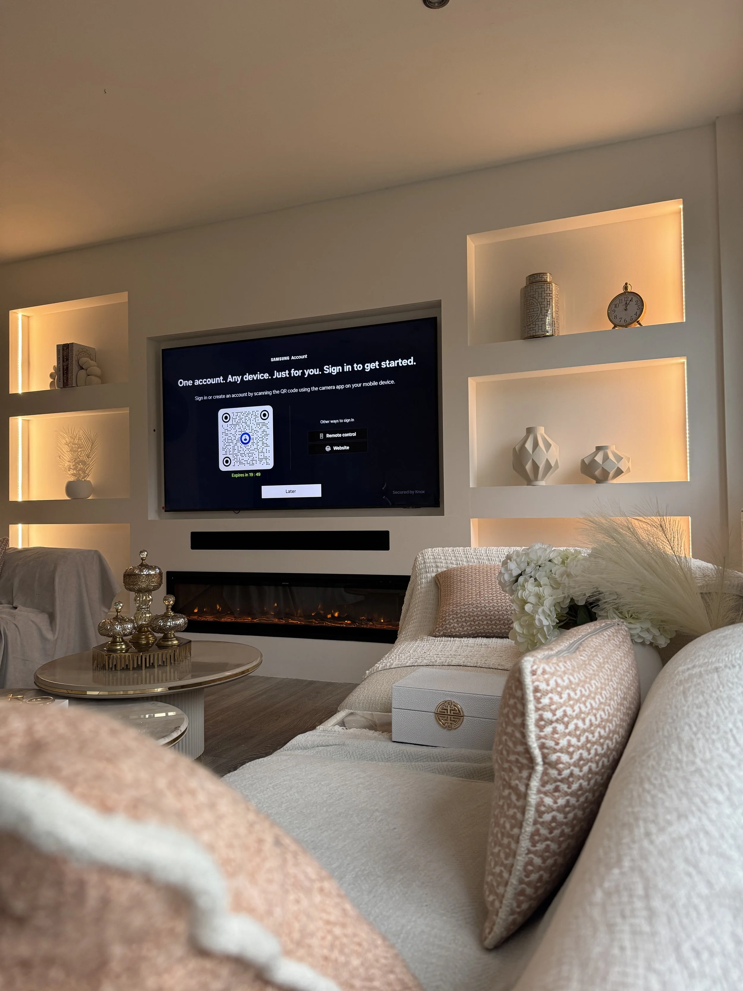 Living room with a wall-mounted television displaying a sign-in screen, a modern electric fireplace below, decorative shelves with vases and clocks, a white sofa with pink cushions, flowers, and a coffee table with decorative items.
