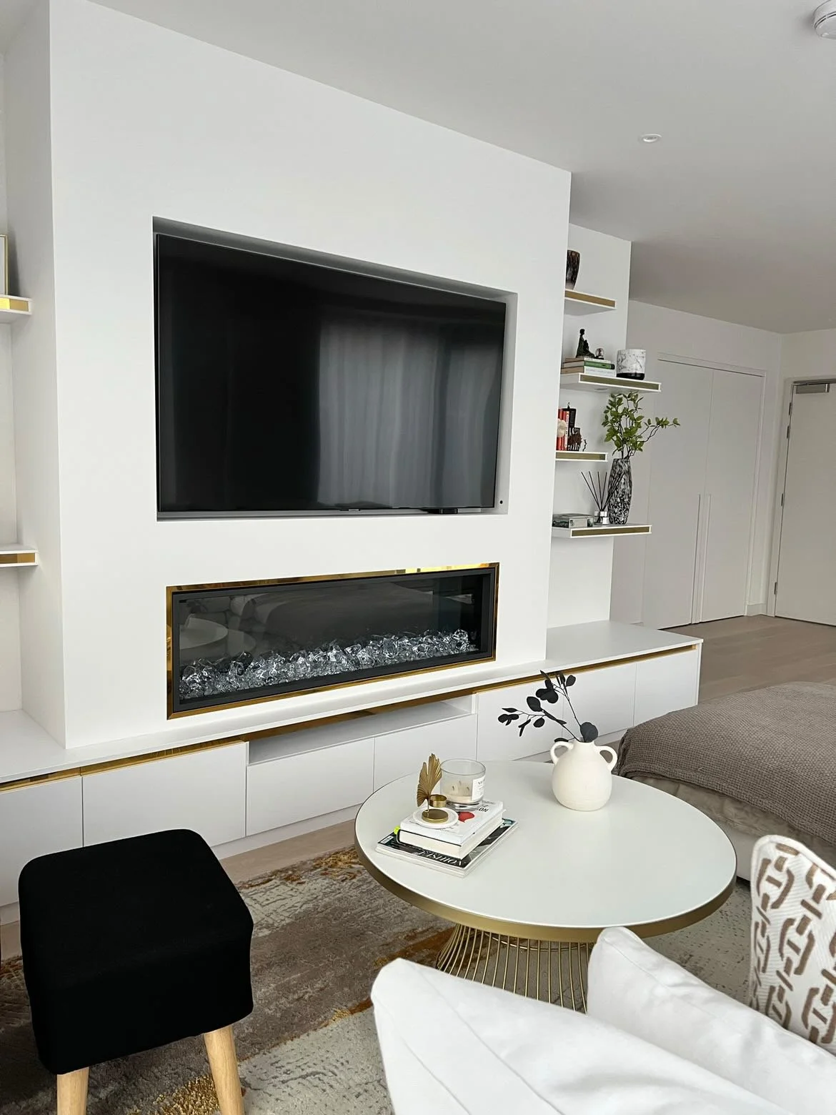 Modern living room with a white entertainment wall, large flat-screen TV, gas fireplace with glass stones, a white round coffee table with decorative items, and a sitting area with various chairs and a bed.
