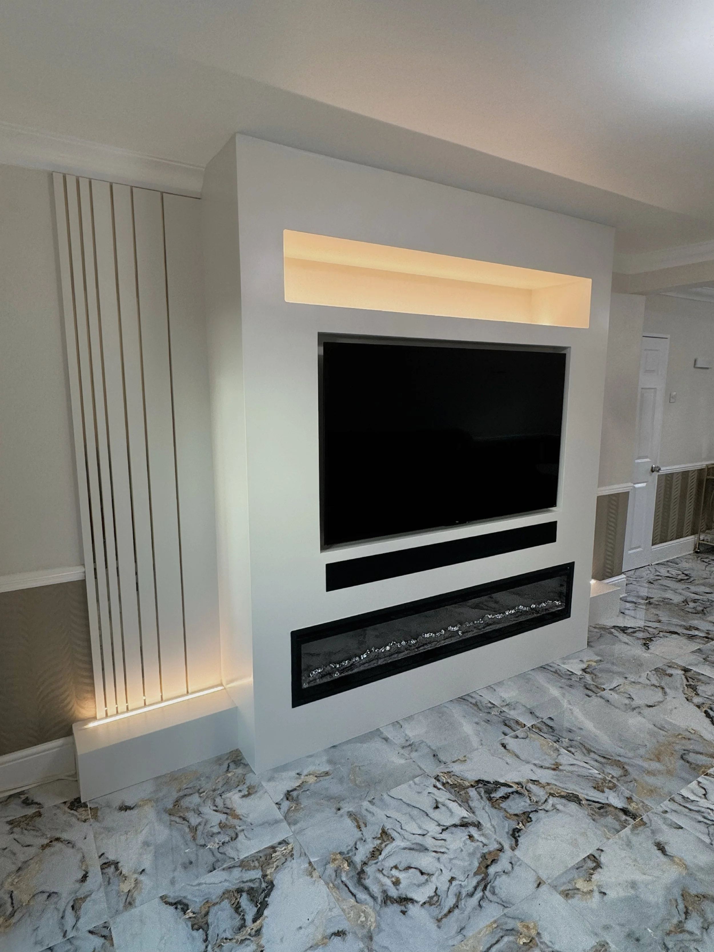 Modern white entertainment center with a mounted flat-screen TV, an illuminated rectangular niche above, and a linear fireplace below, on a marble floor in a contemporary living room.