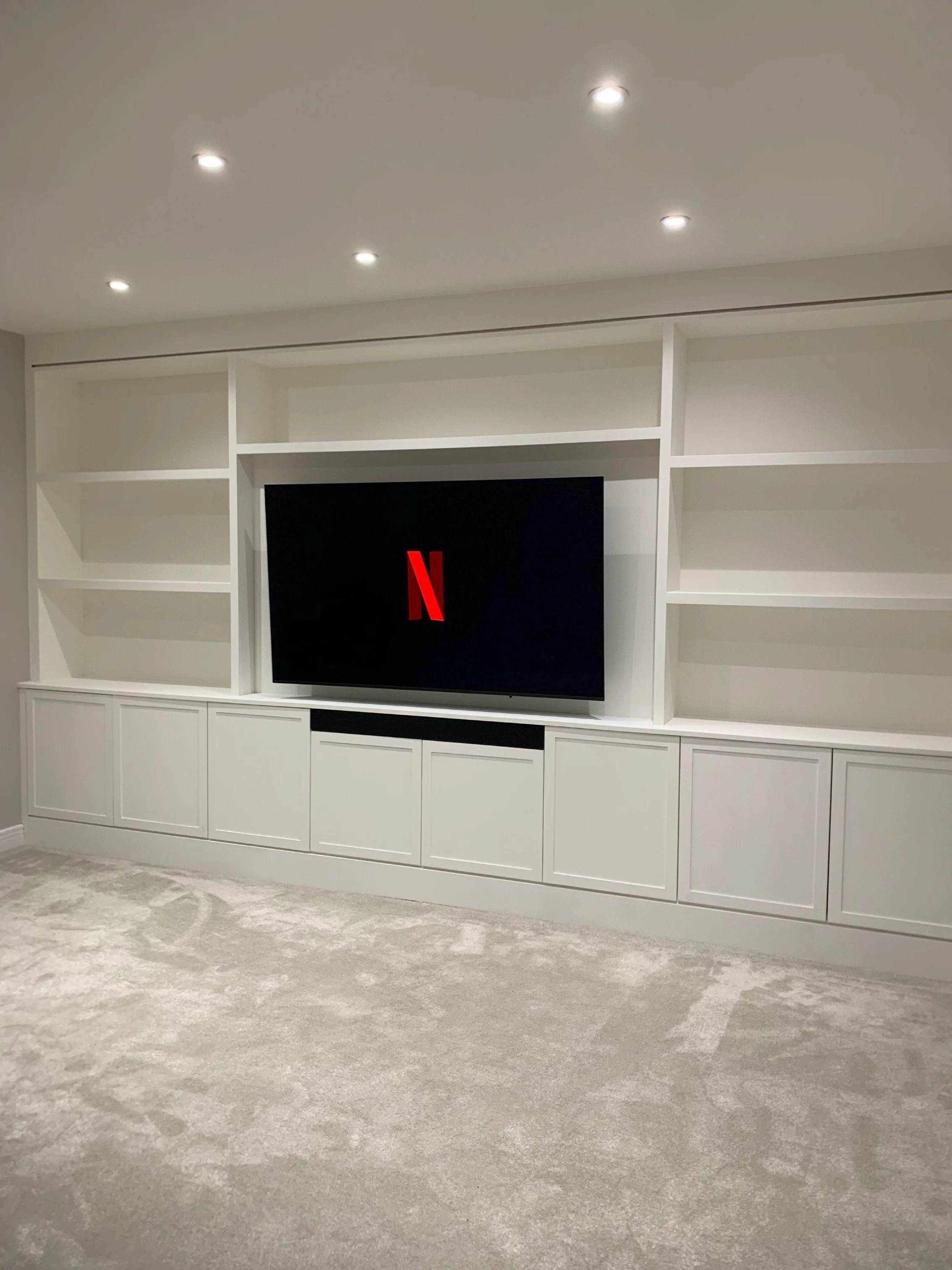 Living room with white built-in shelves, a large flat-screen TV displaying the Netflix logo, and beige carpeted floor.