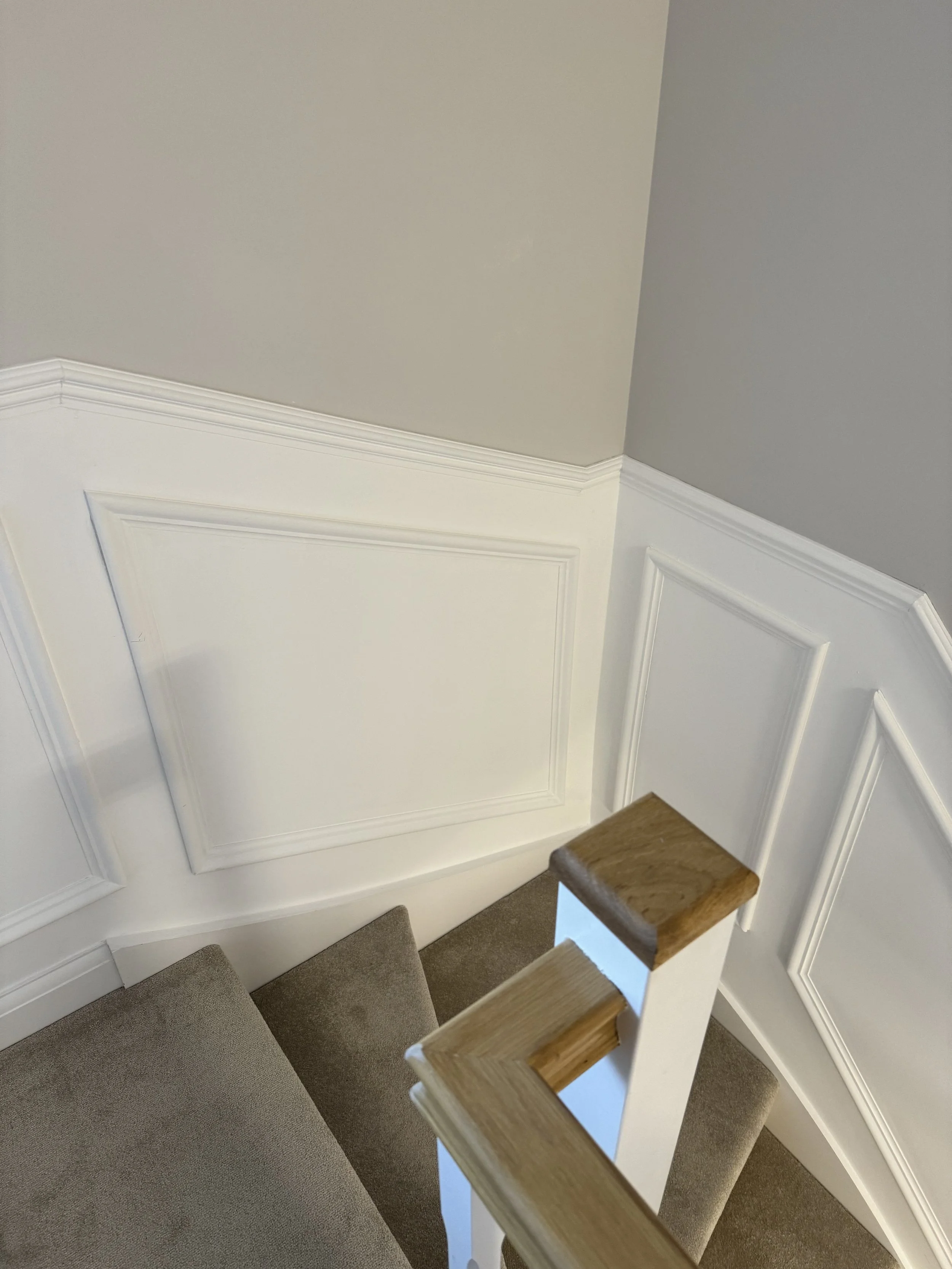 Top view of a staircase with beige carpeted steps and a white wooden handrail with a wooden cap, next to white panel wainscoting on the wall and a gray painted wall.