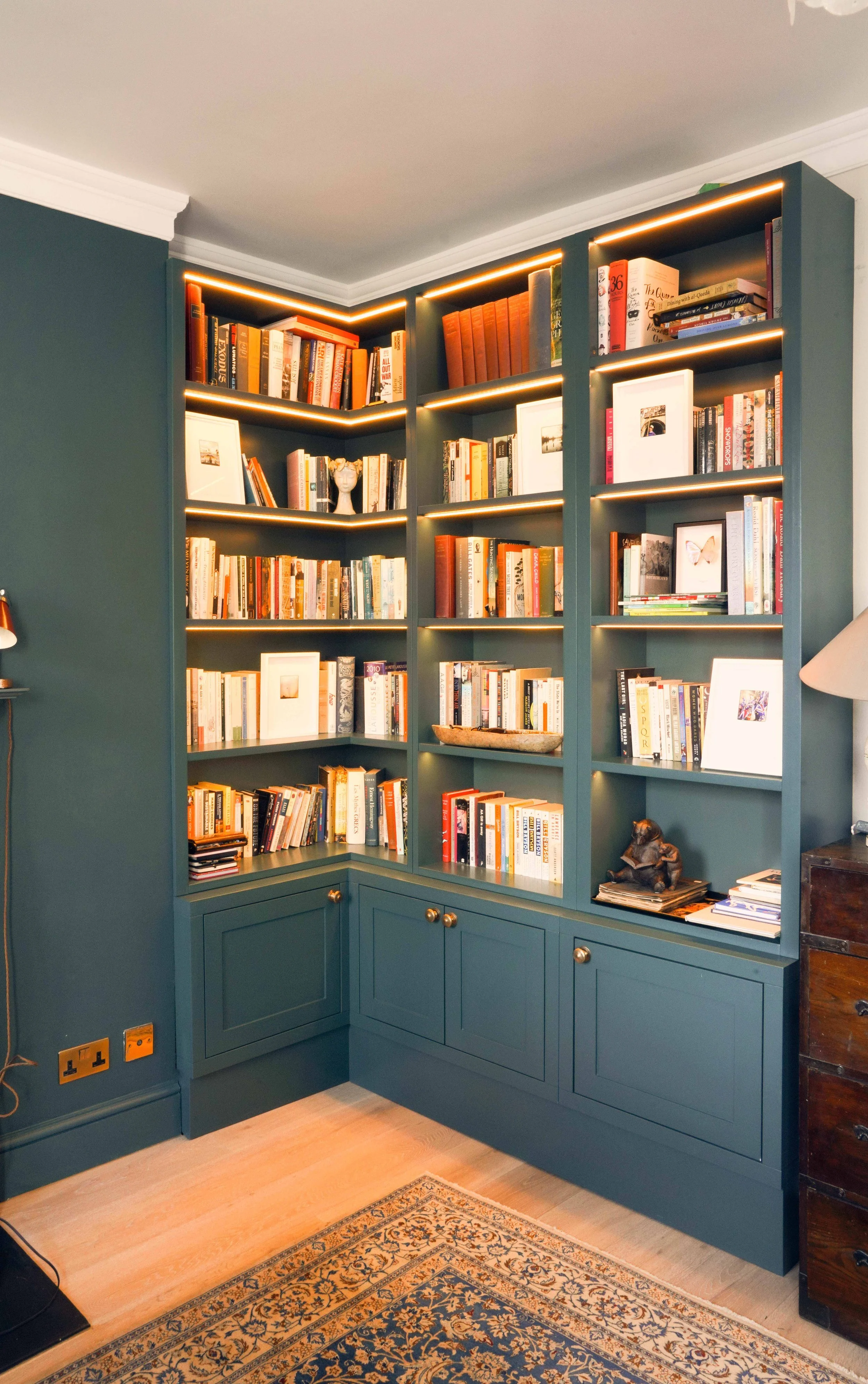 A corner bookshelf filled with books and framed photos, with built-in lighting and gold knobs, against a teal wall and beige carpeted floor.