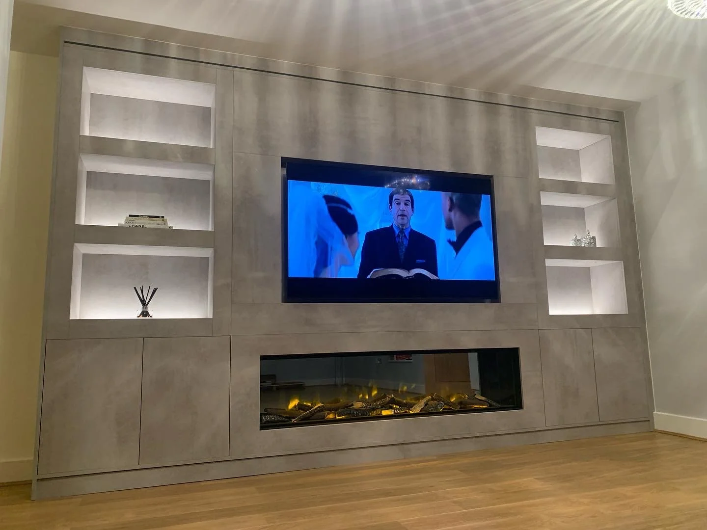 Living room entertainment system with flat-screen TV displaying a scene with three men, built-in shelves with books and decorative items, and an electric fireplace below the TV, all against a gray wall with warm wooden flooring.