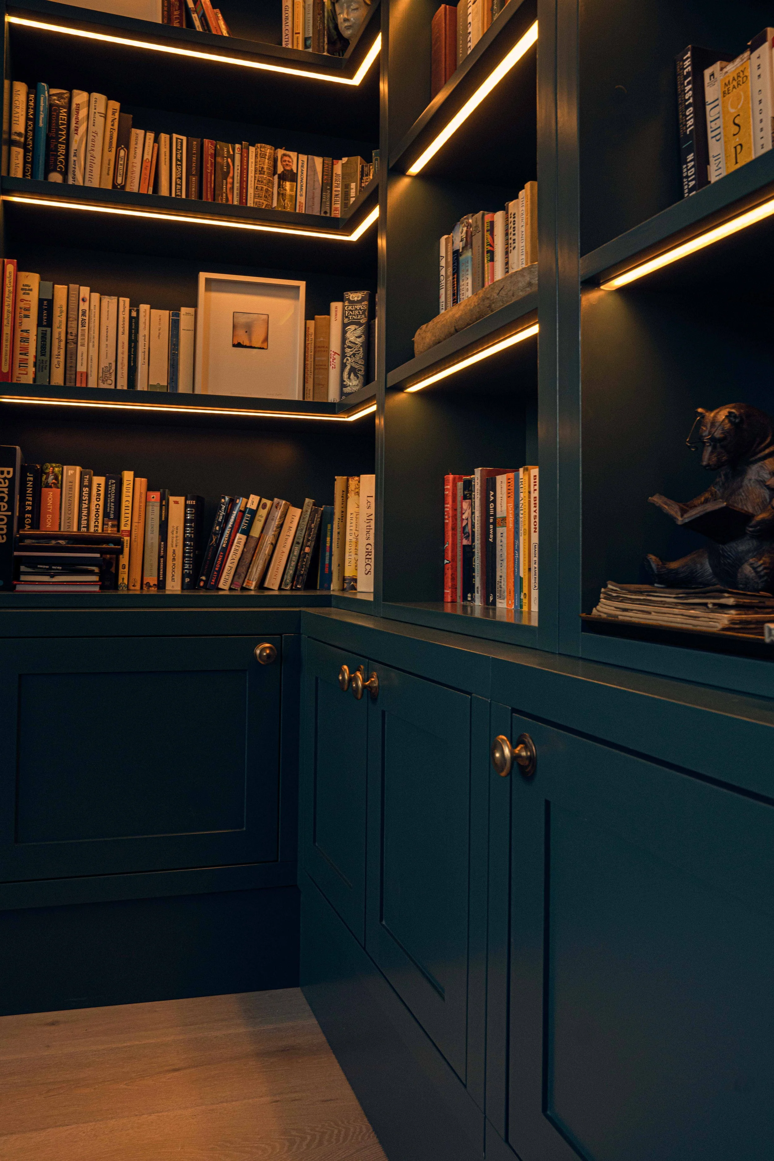 A dark green bookshelf filled with books, decorative objects, and artwork, with integrated lighting highlighting the items and a wooden floor visible at the bottom.