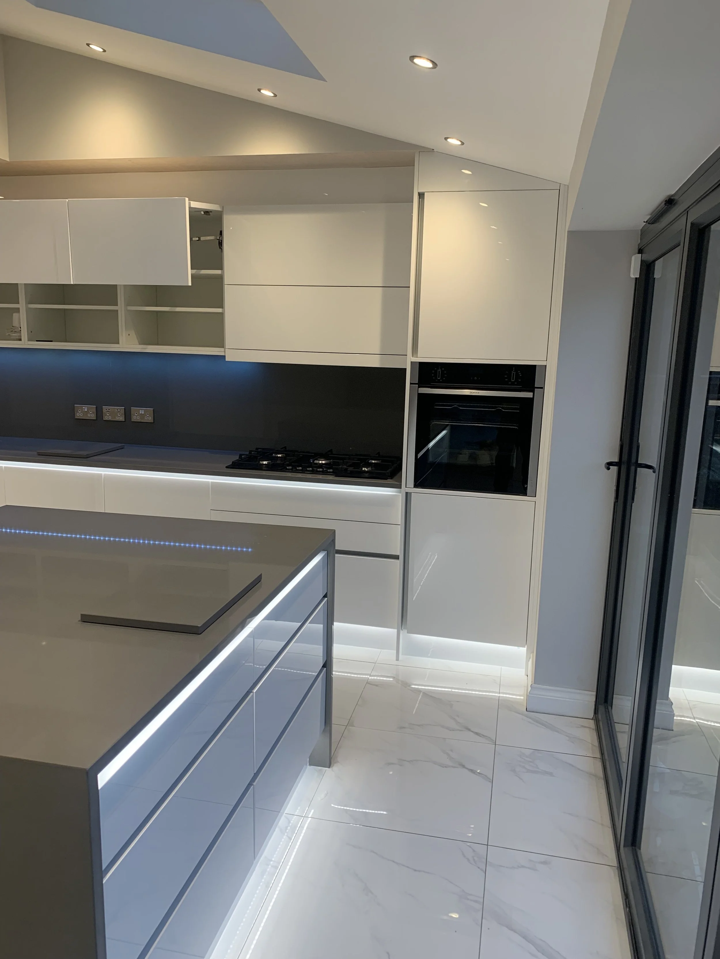 Modern kitchen with white cabinets, a built-in oven, a cooking stove, and glossy white floor tiles. There is a kitchen island with LED lighting underneath and a sliding door on the right.