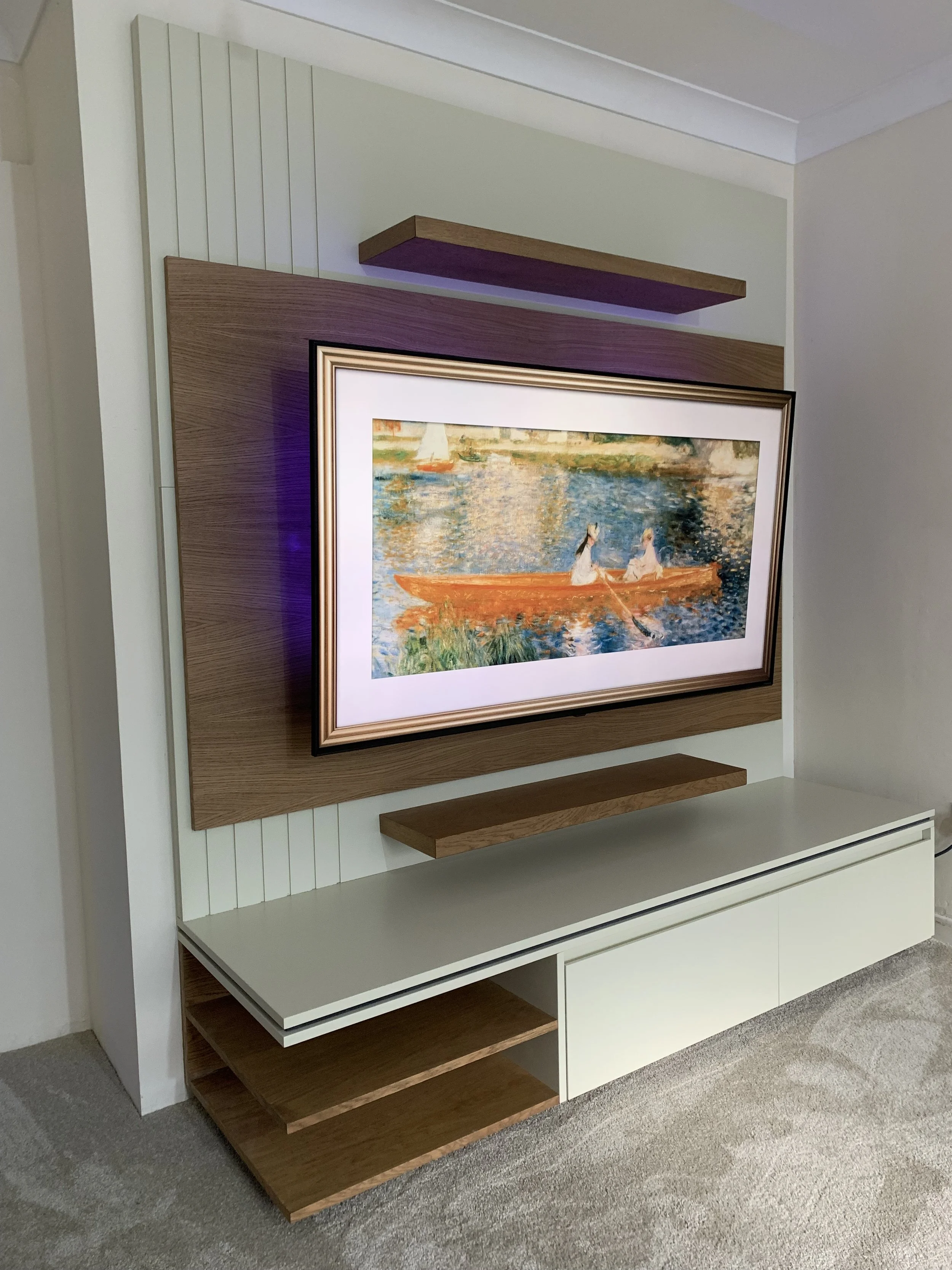 Modern living room with a wall-mounted, framed landscape painting of two women in a boat on a river, with wooden shelves and a white entertainment unit.