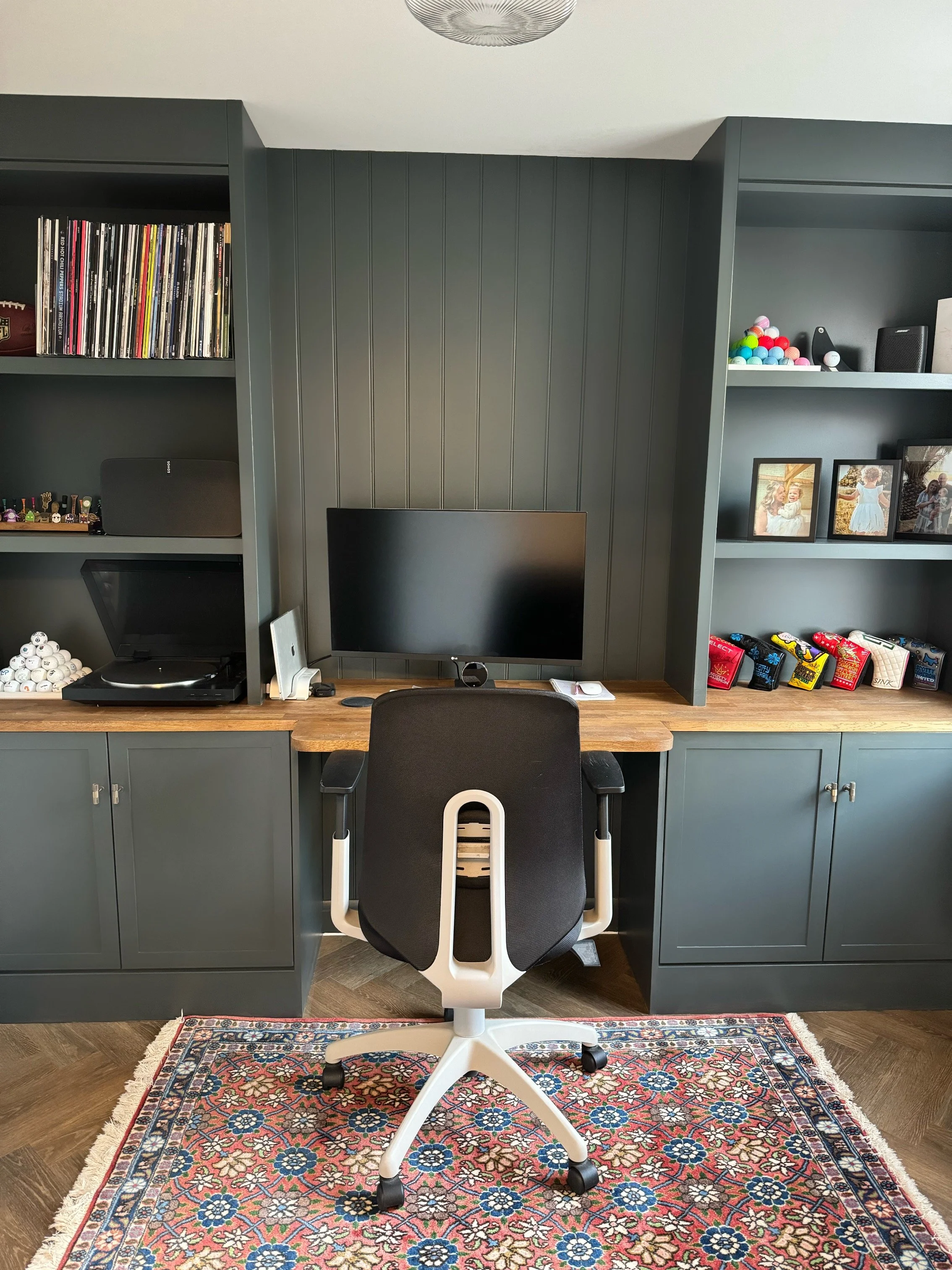 Home office with dark gray cabinetry, a wooden desk, computer monitor, office chair, a patterned rug, and shelves with books, photos, and decorative items.