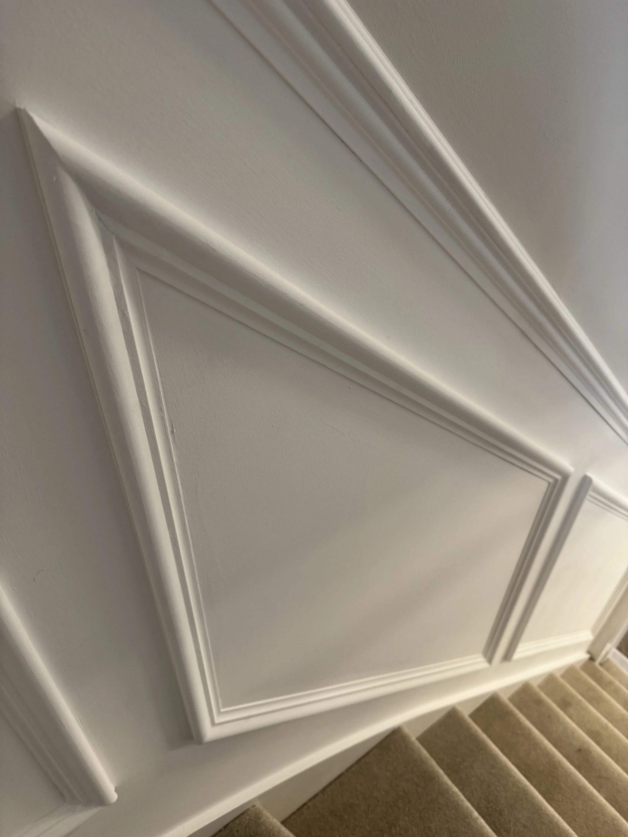 Close-up of decorative white wall paneling with molding, next to beige carpeted stairs.
