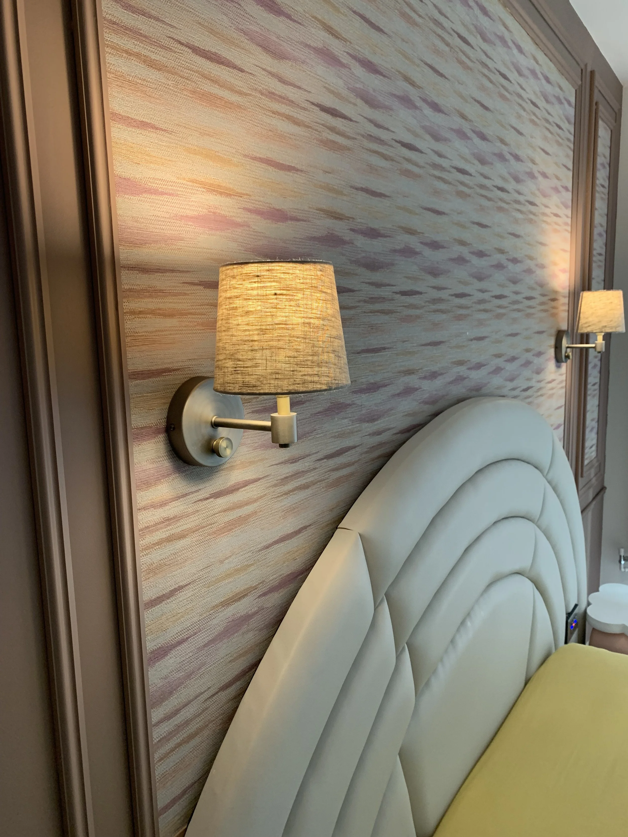 Close-up of a bedroom wall with pink, purple, and beige textured wallpaper, featuring two wall-mounted lamps with beige shades, a cream-colored padded headboard, and a yellow bedspread.