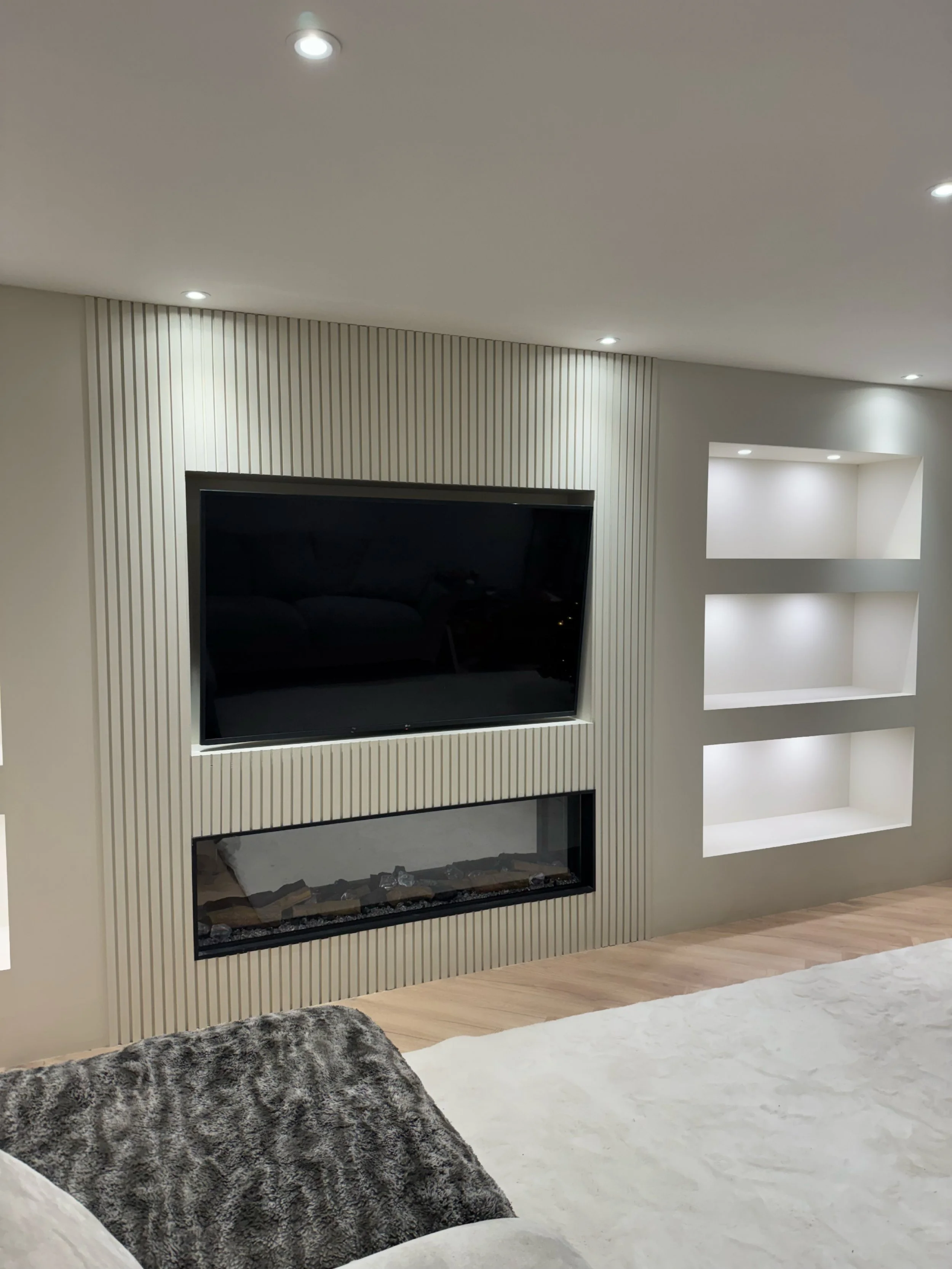 Modern living room with built-in wall features including a flat-screen TV, an electric fireplace, and illuminated niche shelves. The room has recessed ceiling lights, a white carpet, and a plush chair with a fur-like throw.