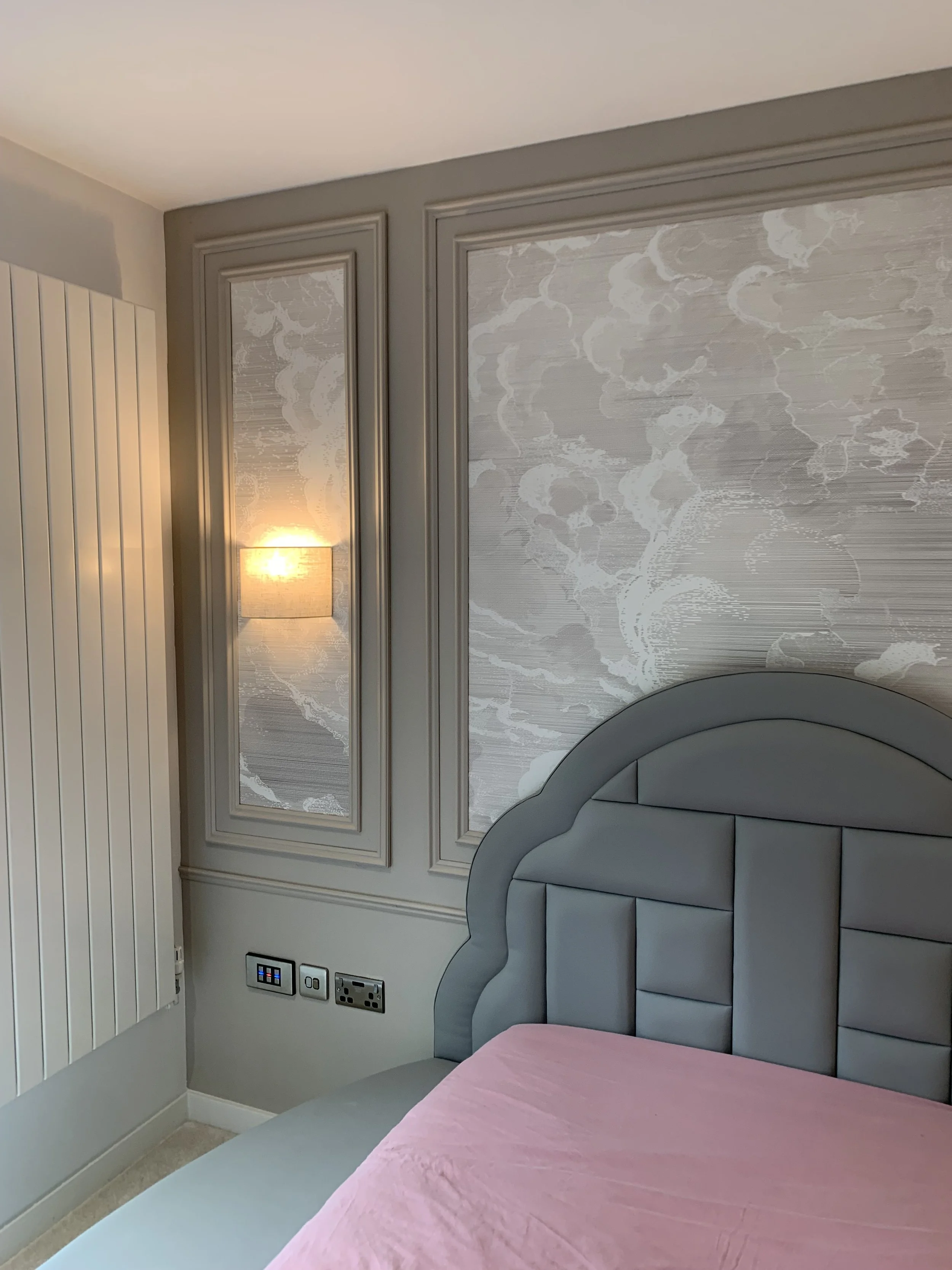 Close-up of a bedroom with gray upholstered headboard, pink bedspread, decorative wall panels, and a wall-mounted lamp.