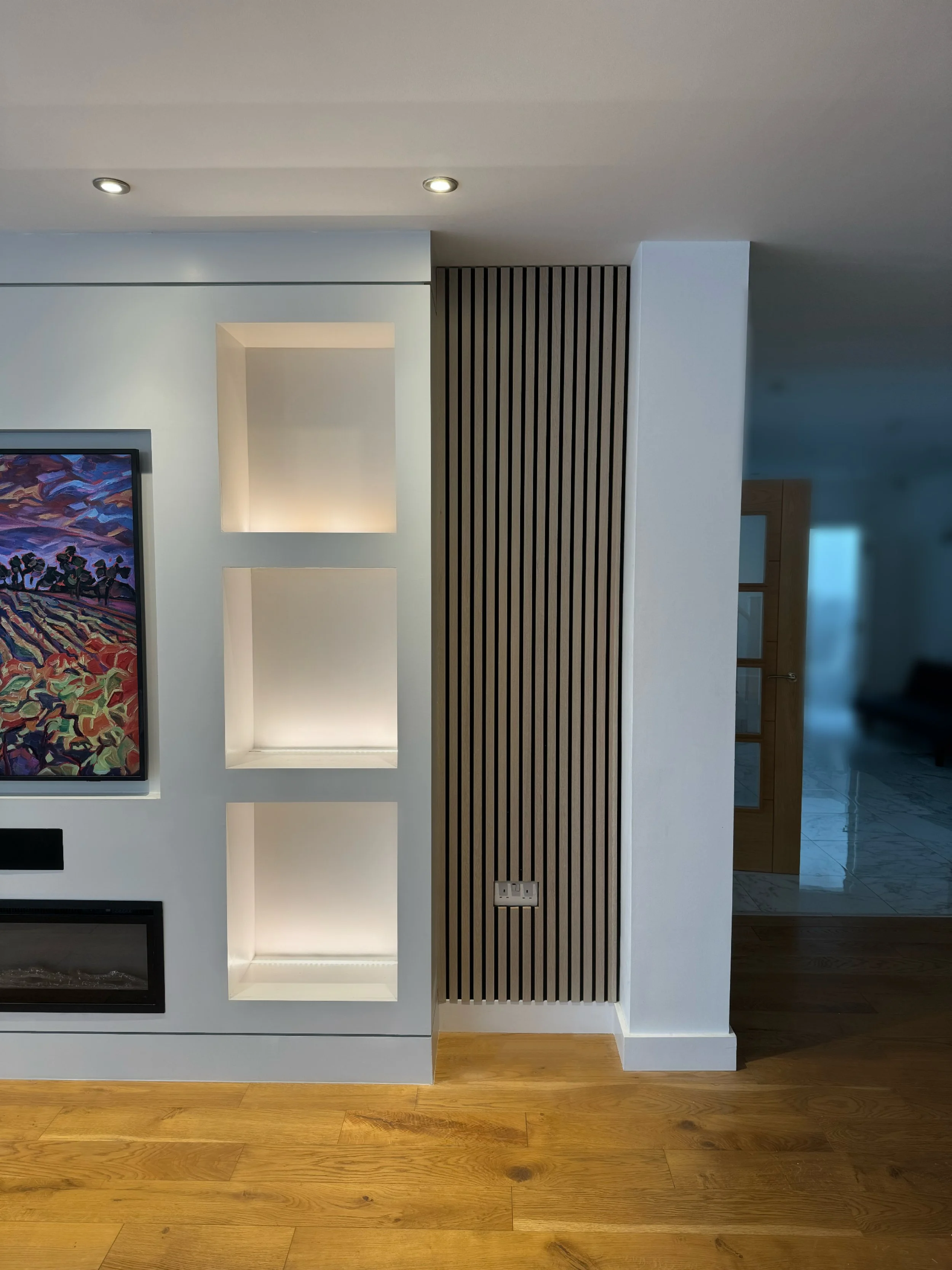 Interior of a modern living room with wooden flooring, white walls, a colorful abstract painting, and built-in illuminated shelves next to a wooden slatted partition.